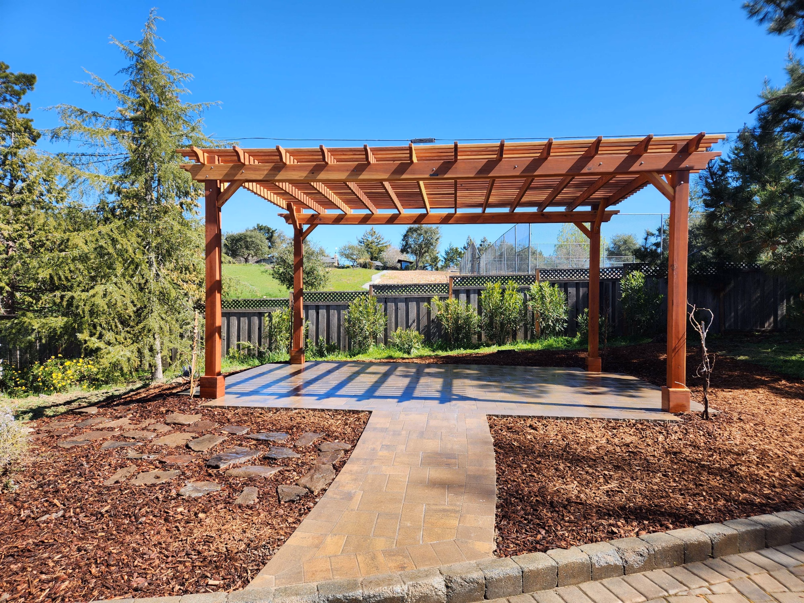 A redwood-stained timber pergola with shaped rafter tails over a paver patio, with a paver walkway and new planting, in Los Gatos