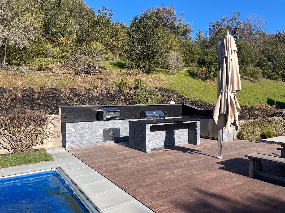 Dark stacked-stone outdoor kitchen with two grills on a composite deck beside a pool, with a hillside behind, in Woodside, CA