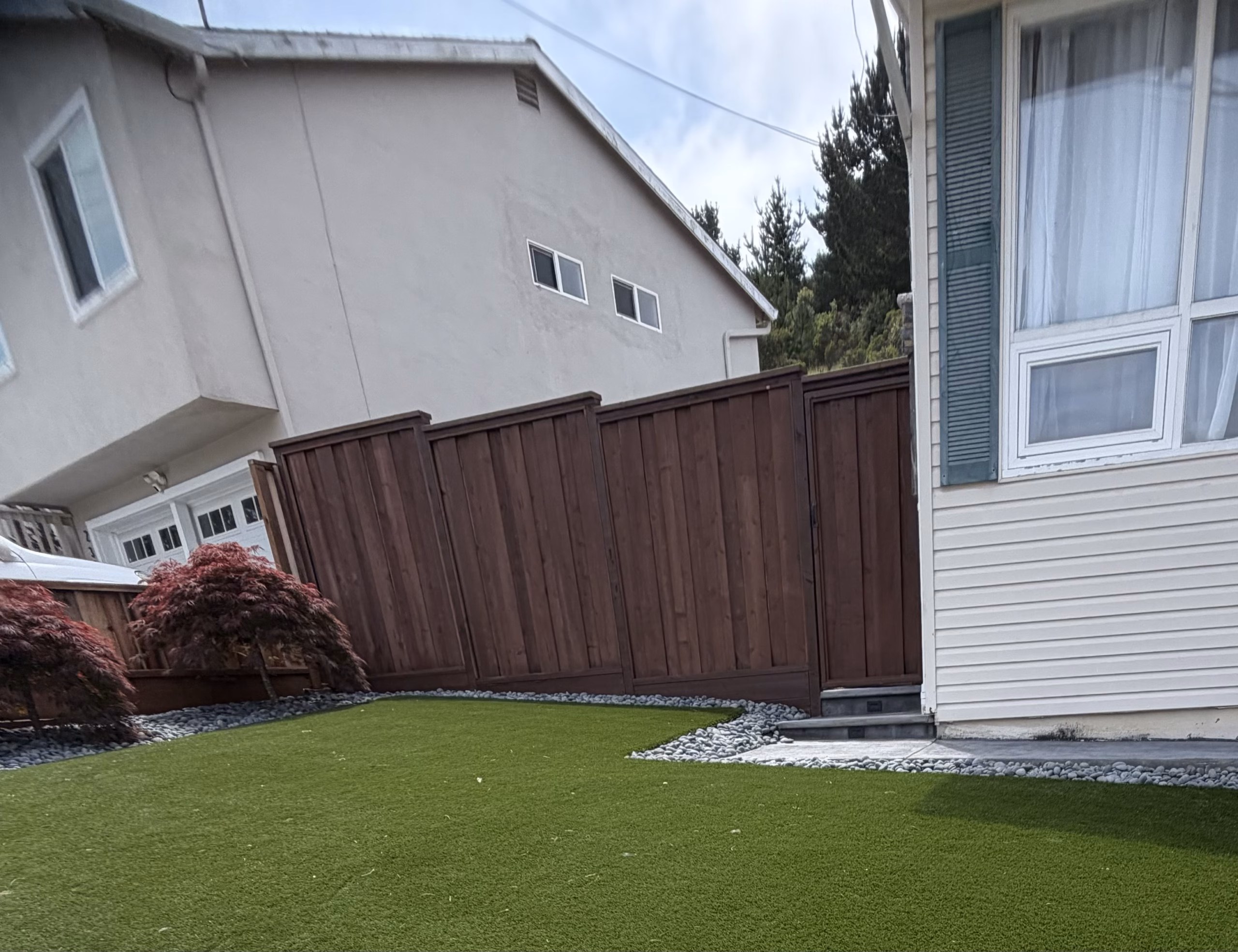 Small artificial-turf lawn beside a two-story house and a wood fence, with a gravel border, in San Bruno, CA
