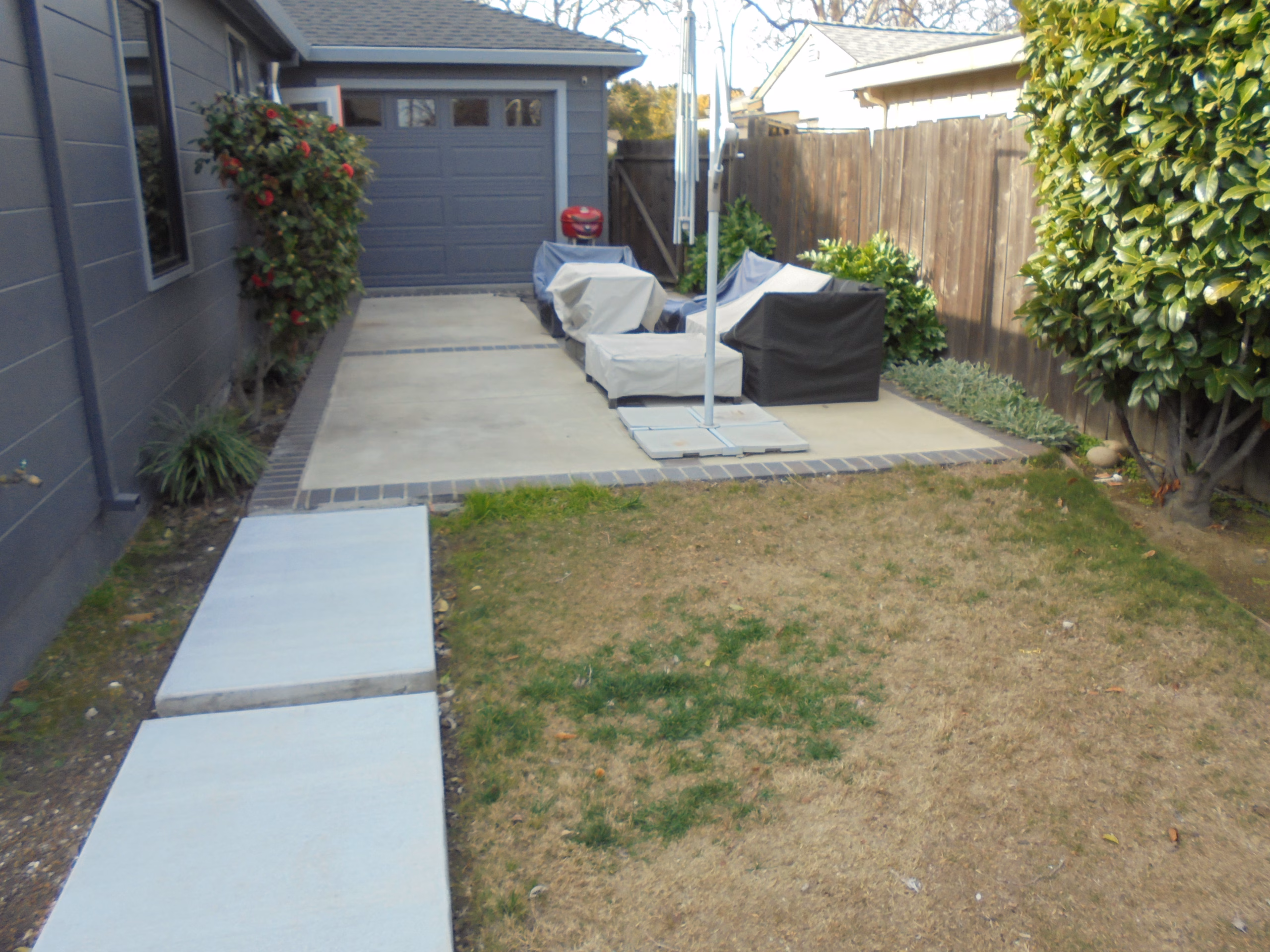 A poured-concrete walkway along the side of a gray house leading to a concrete patio with lounge furniture, beside a lawn, in Redwood City