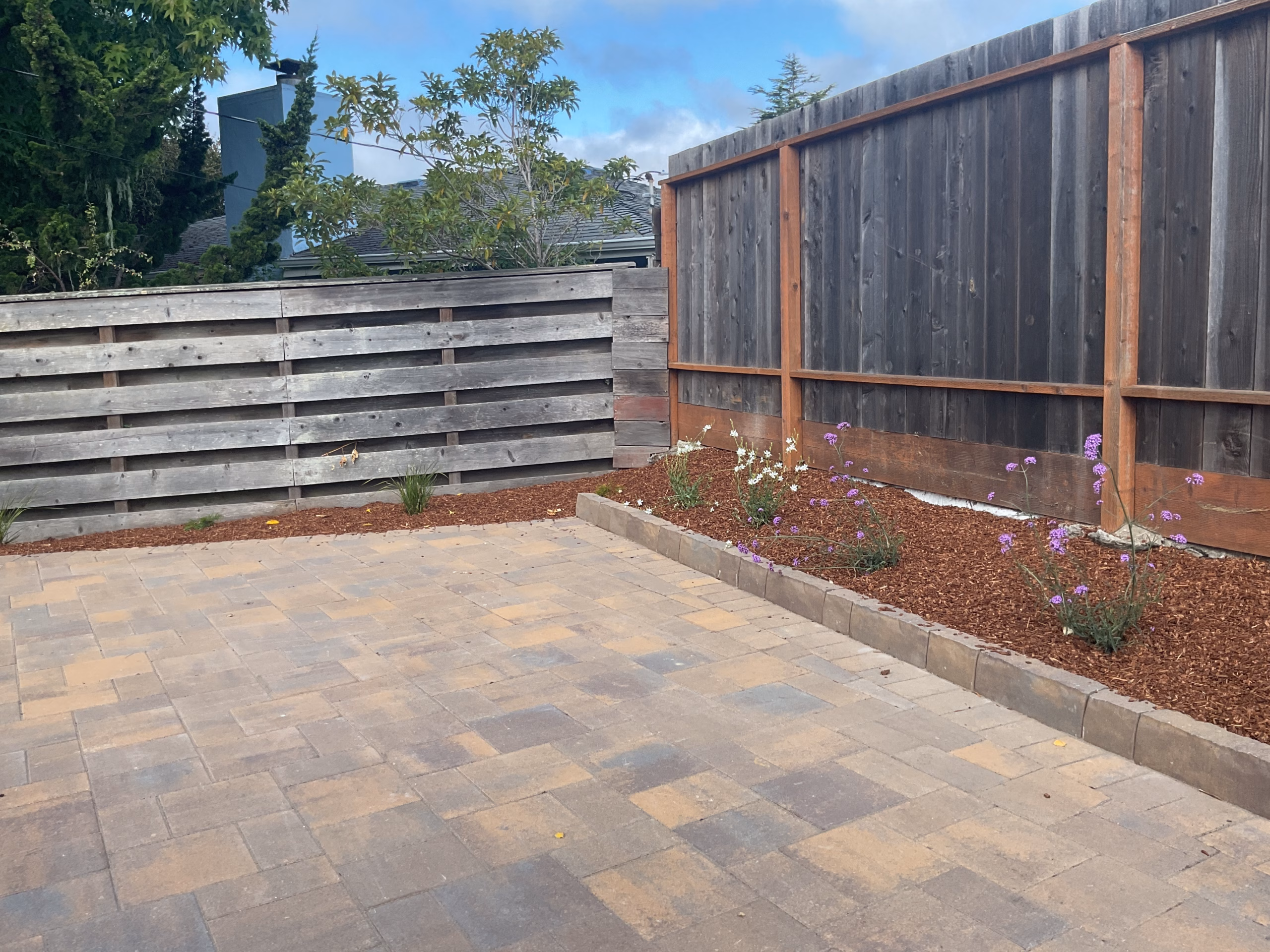 A tan-and-gray interlocking paver patio with mulched planting beds along a new wood fence, in Millbrae