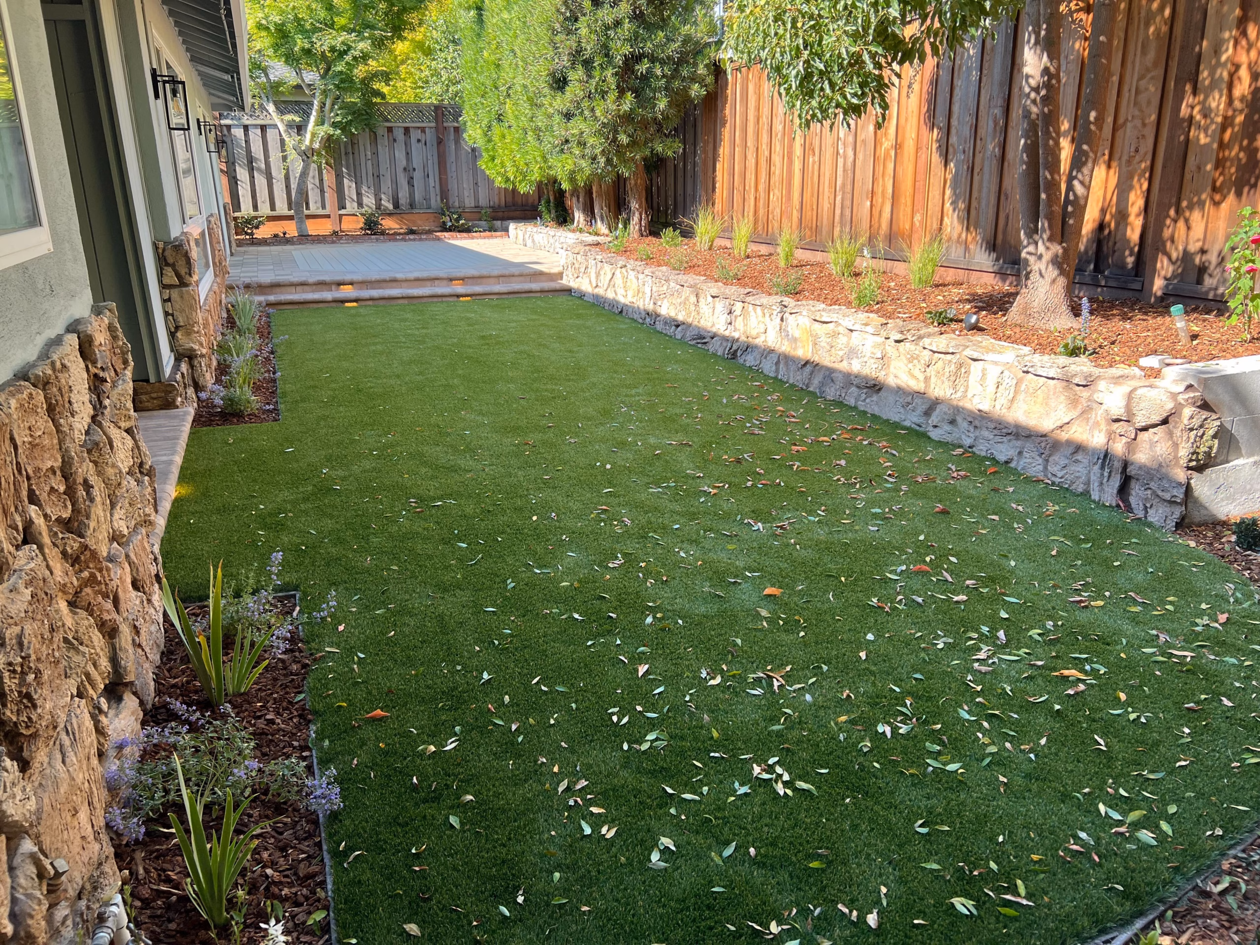 Long lawn panel edged with a stacked-stone border and mulched planting beds along a redwood fence in San Carlos, CA
