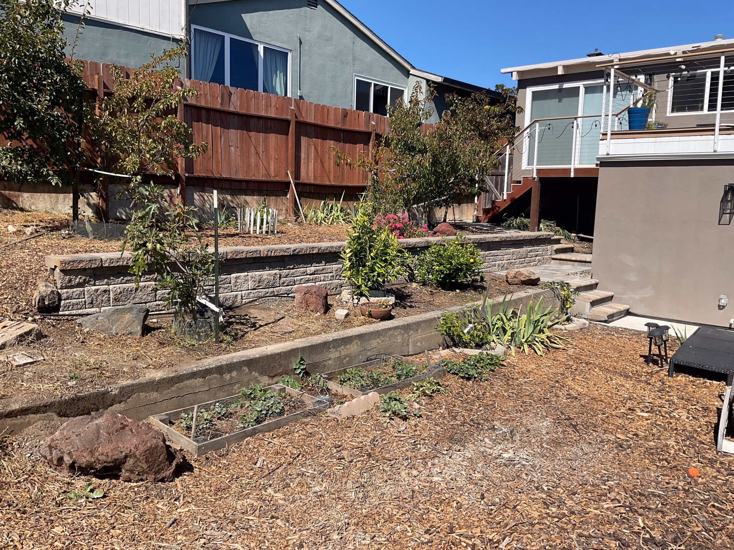 Block retaining wall and a mulched planting bed below a house deck, with a paver step, in San Bruno, CA