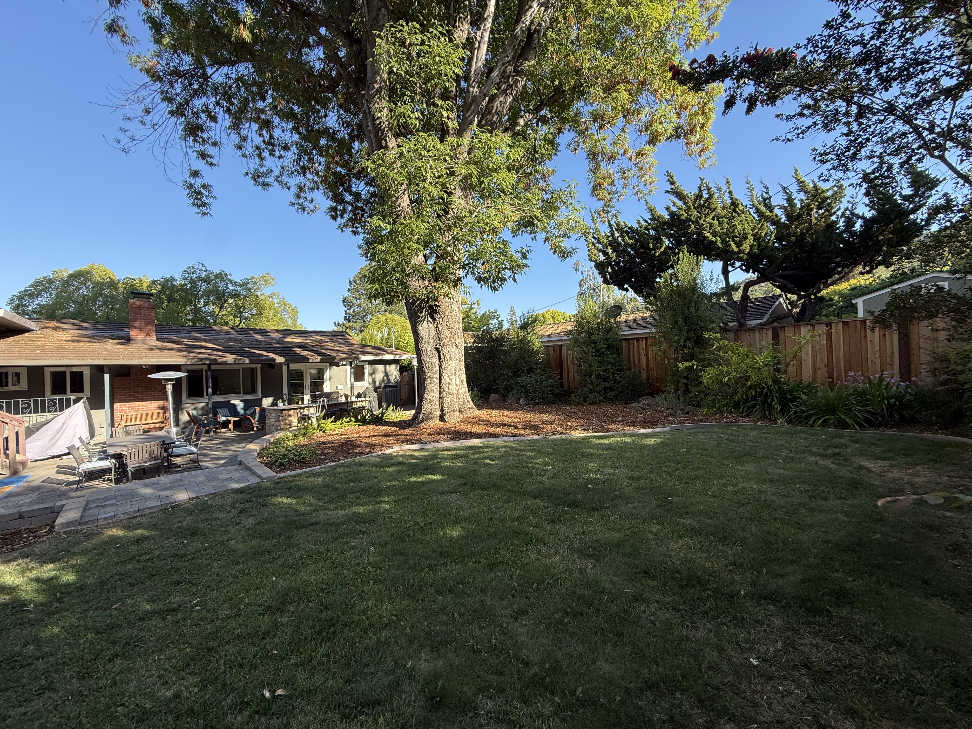 Original-state wide backyard in good light showing a single-story house, a small existing patio with scattered furniture,...