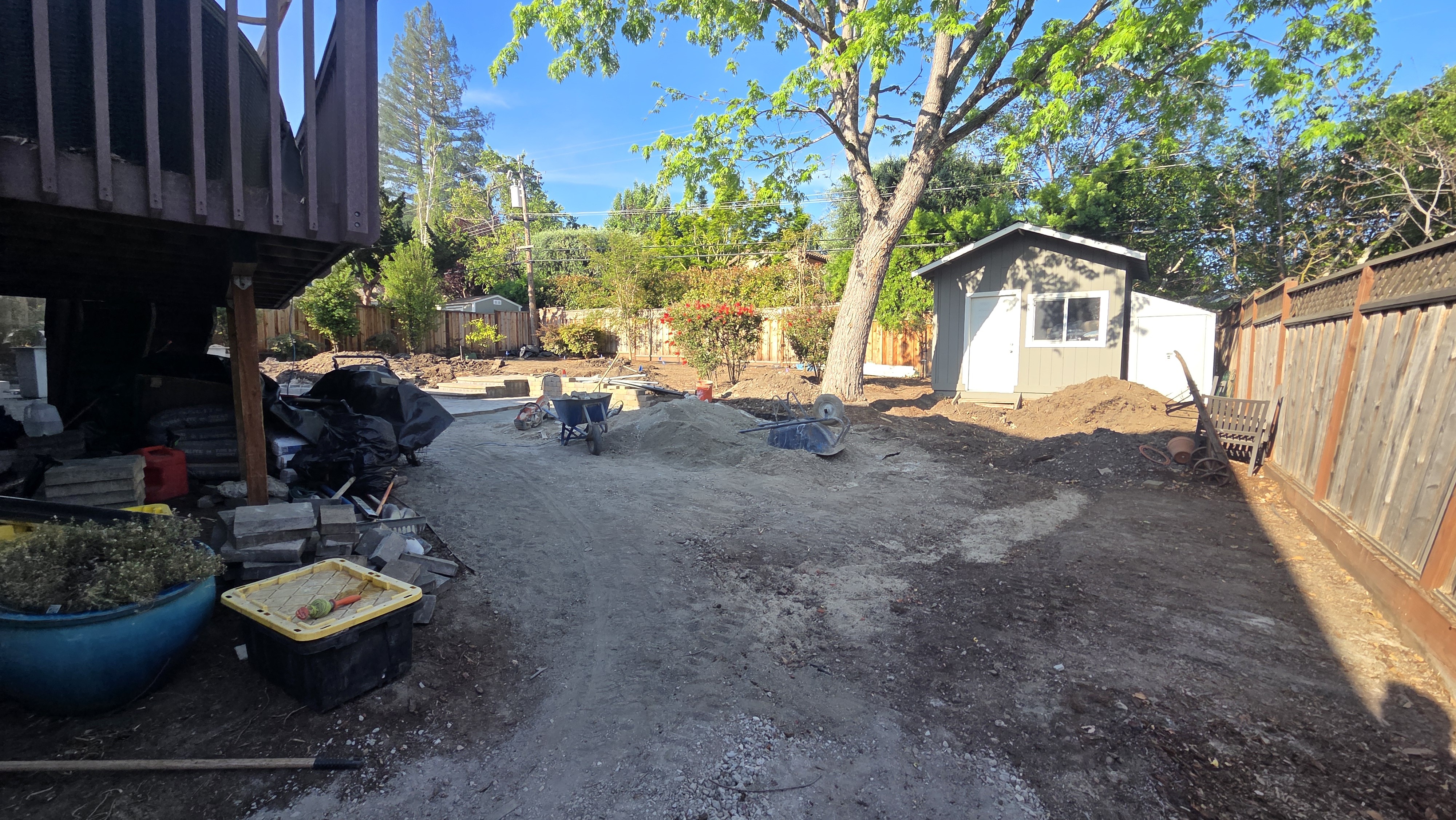 Early base-prep view of the cleared dirt yard with a wheelbarrow, debris piles and bins staged under the deck and the...
