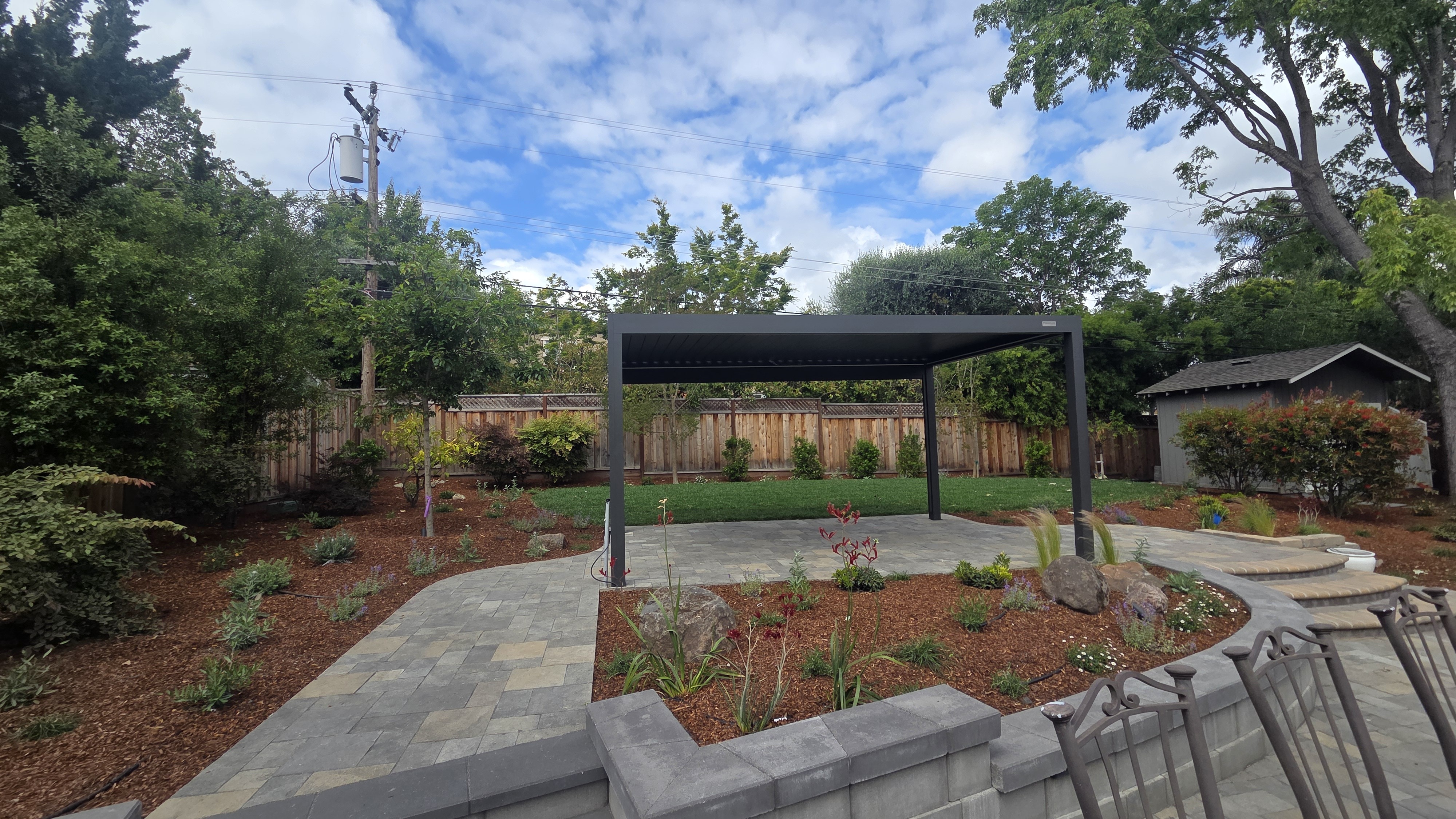 Finished backyard landscape with a modern black pergola centered over fresh planting beds and sod lawn, connected by...