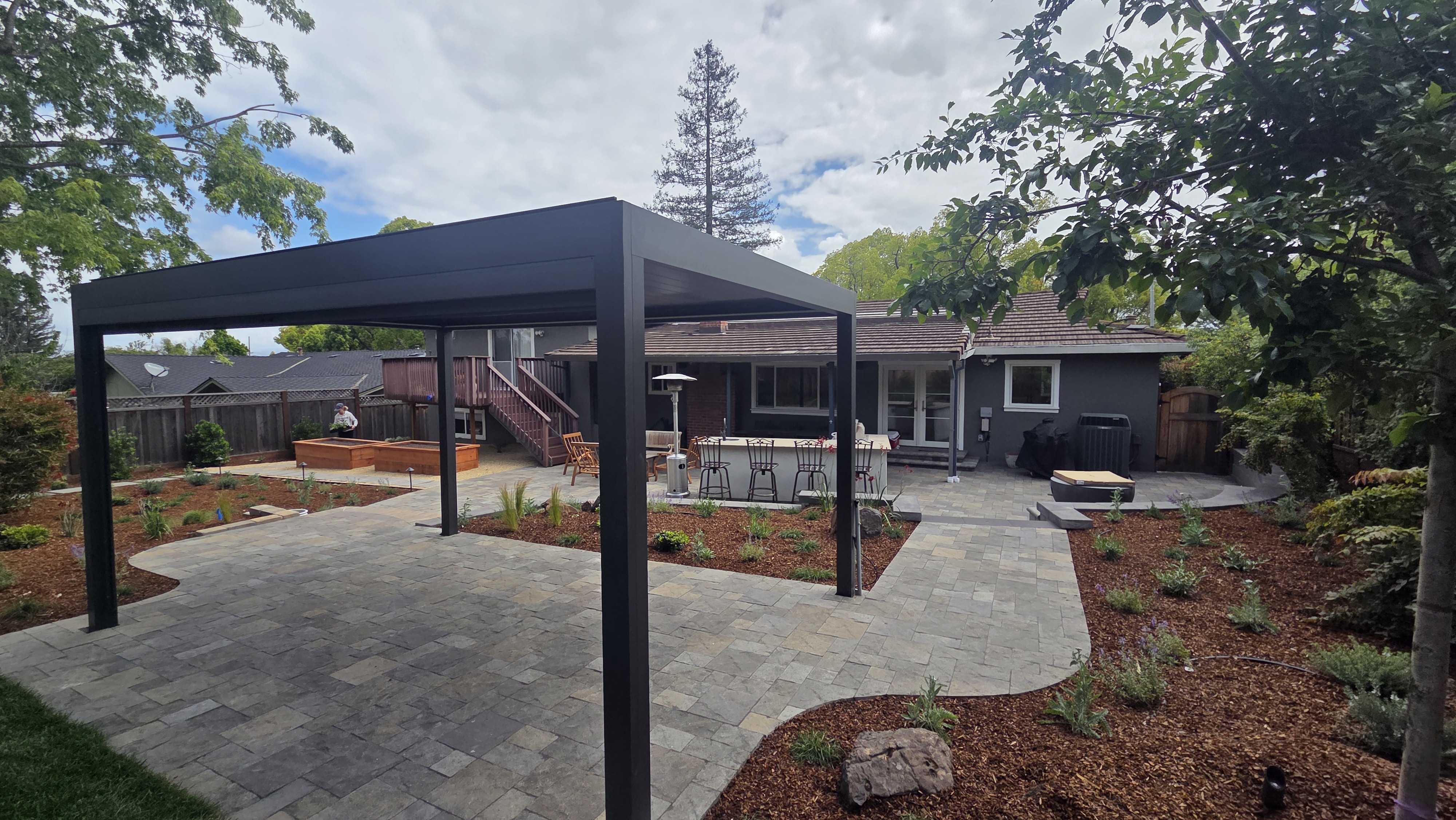 Finished wide view of the large black pergola over a patterned paver patio with bar seating and fire bowl set against the...