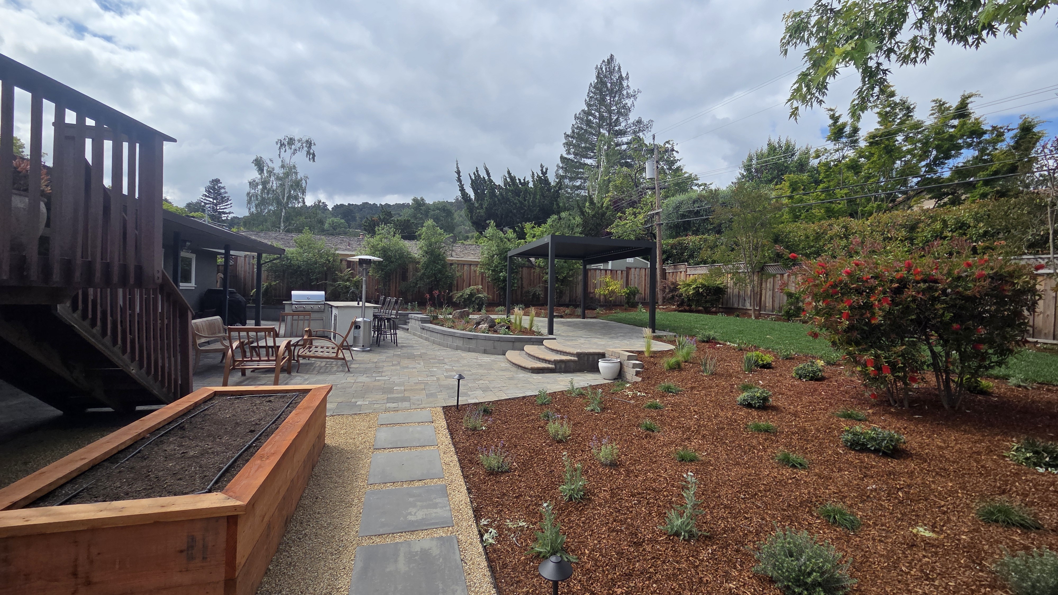 Finished backyard showing newly planted mulch beds and a bluestone path leading to a circular paver patio with a black...
