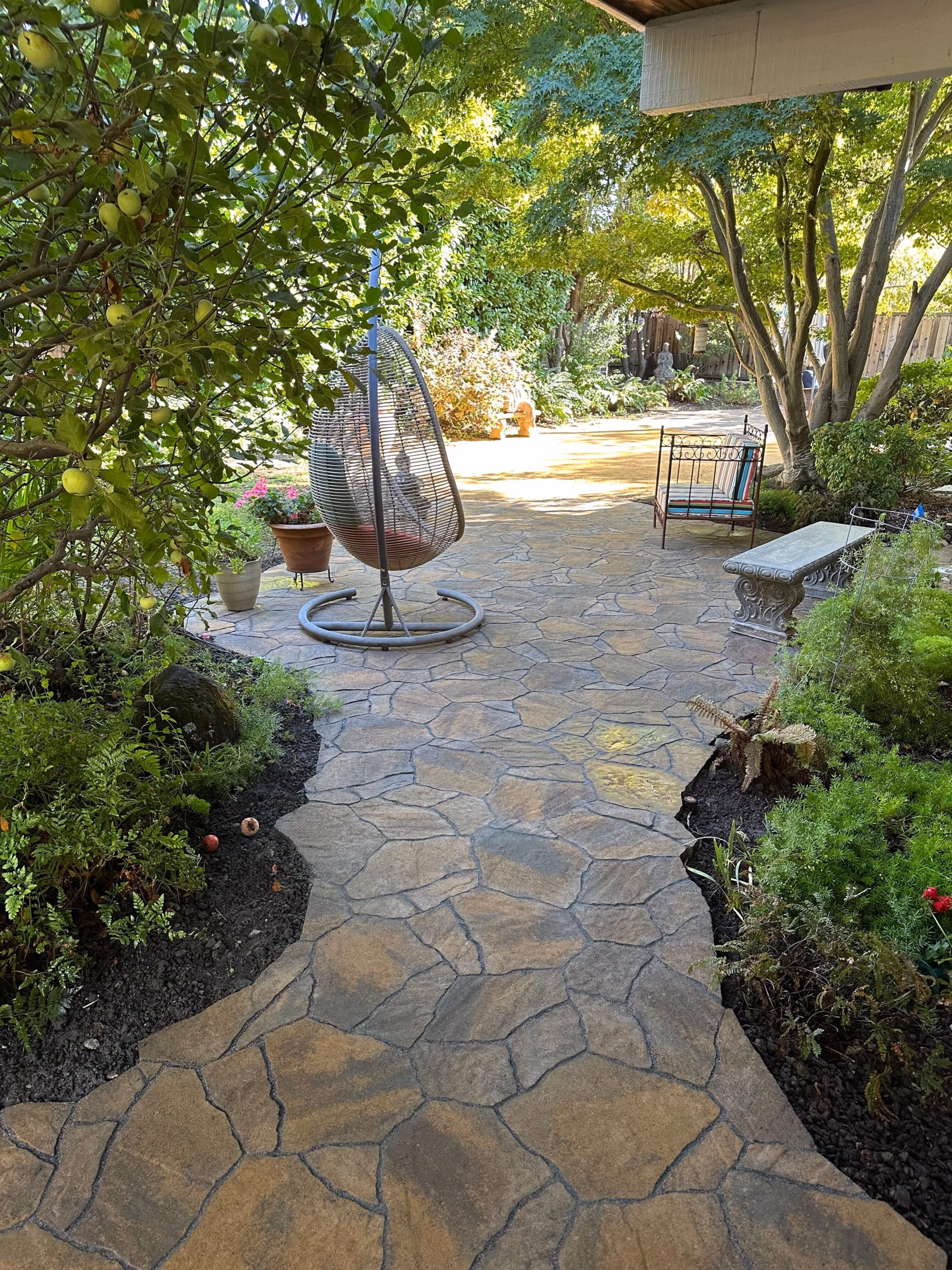 Flagstone-pattern paver path curving through a planted Palo Alto garden past a hanging chair and stone bench under mature trees.