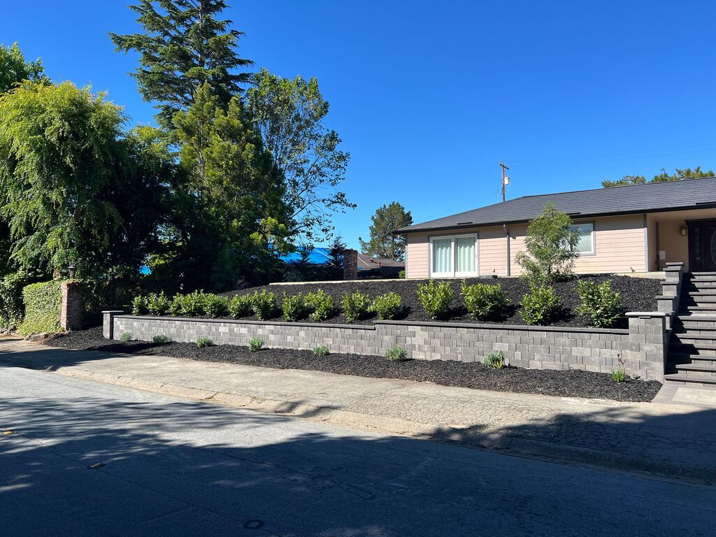 Long gray segmental-block retaining wall along the street with a freshly mulched terrace of new shrubs.