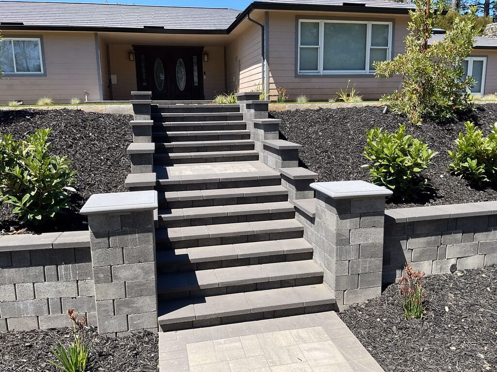 Wide gray paver staircase rising between capped segmental-block pilasters to the front door, flanked by mulched shrub beds.