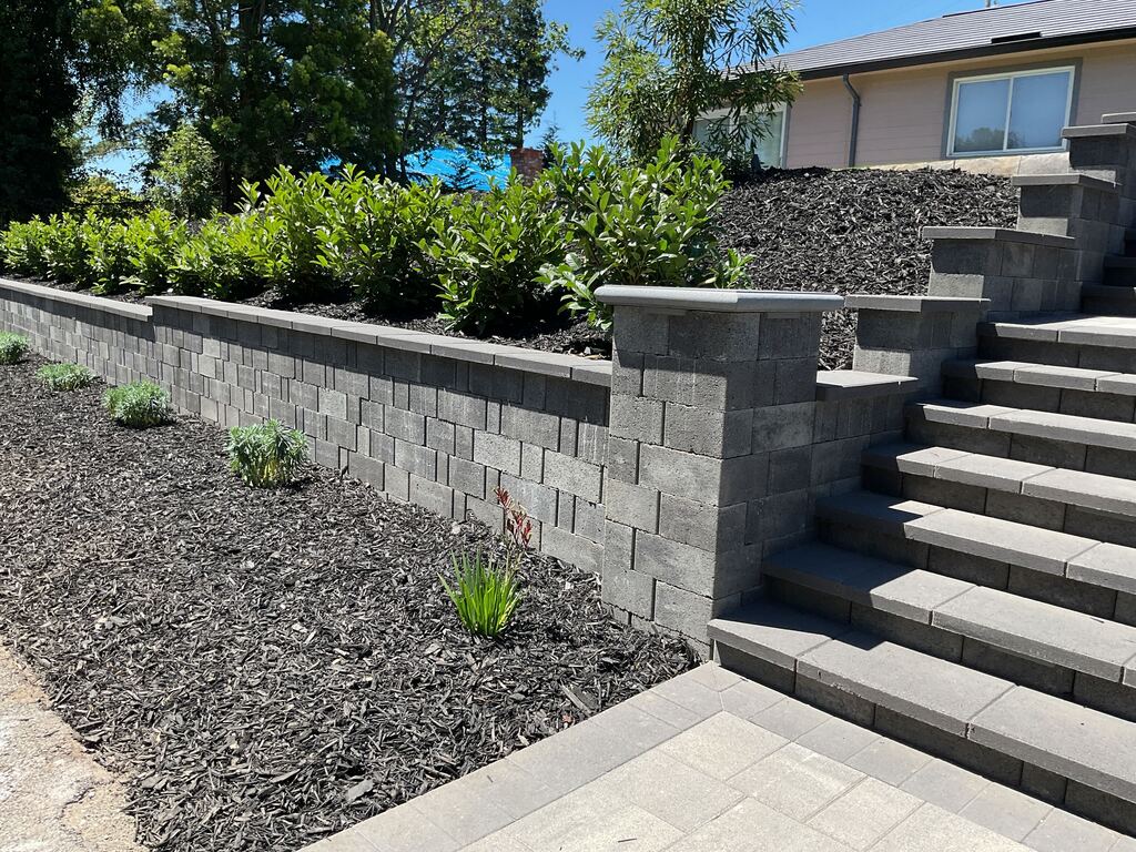 Capped segmental-block retaining wall and pilaster meeting the paver entry steps above a mulched planting bed.
