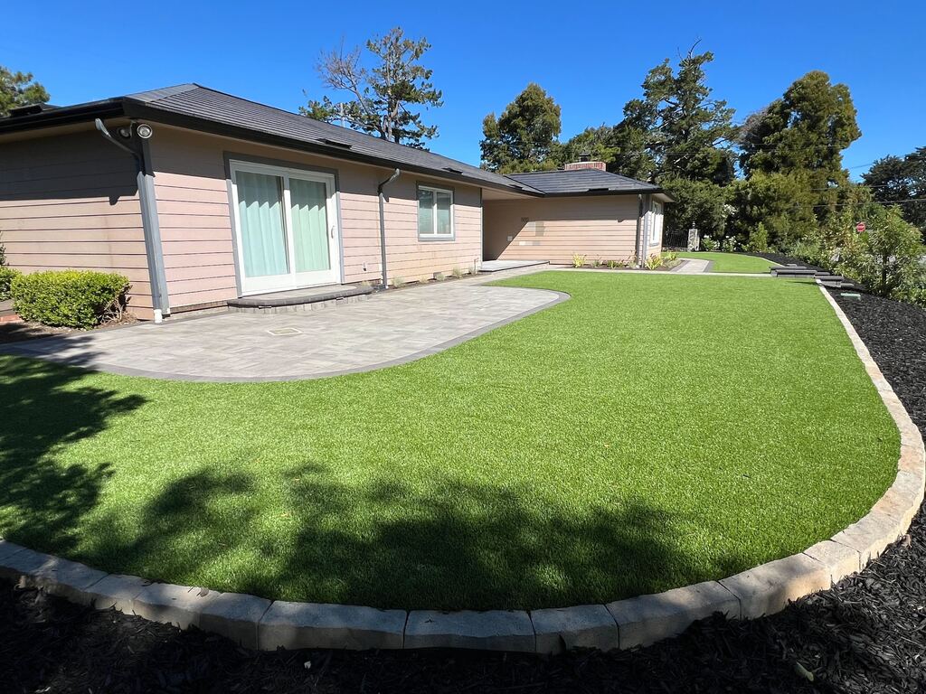 Curved artificial-turf lawn wrapping the house corner with a paver patio strip and a stone-capped border.