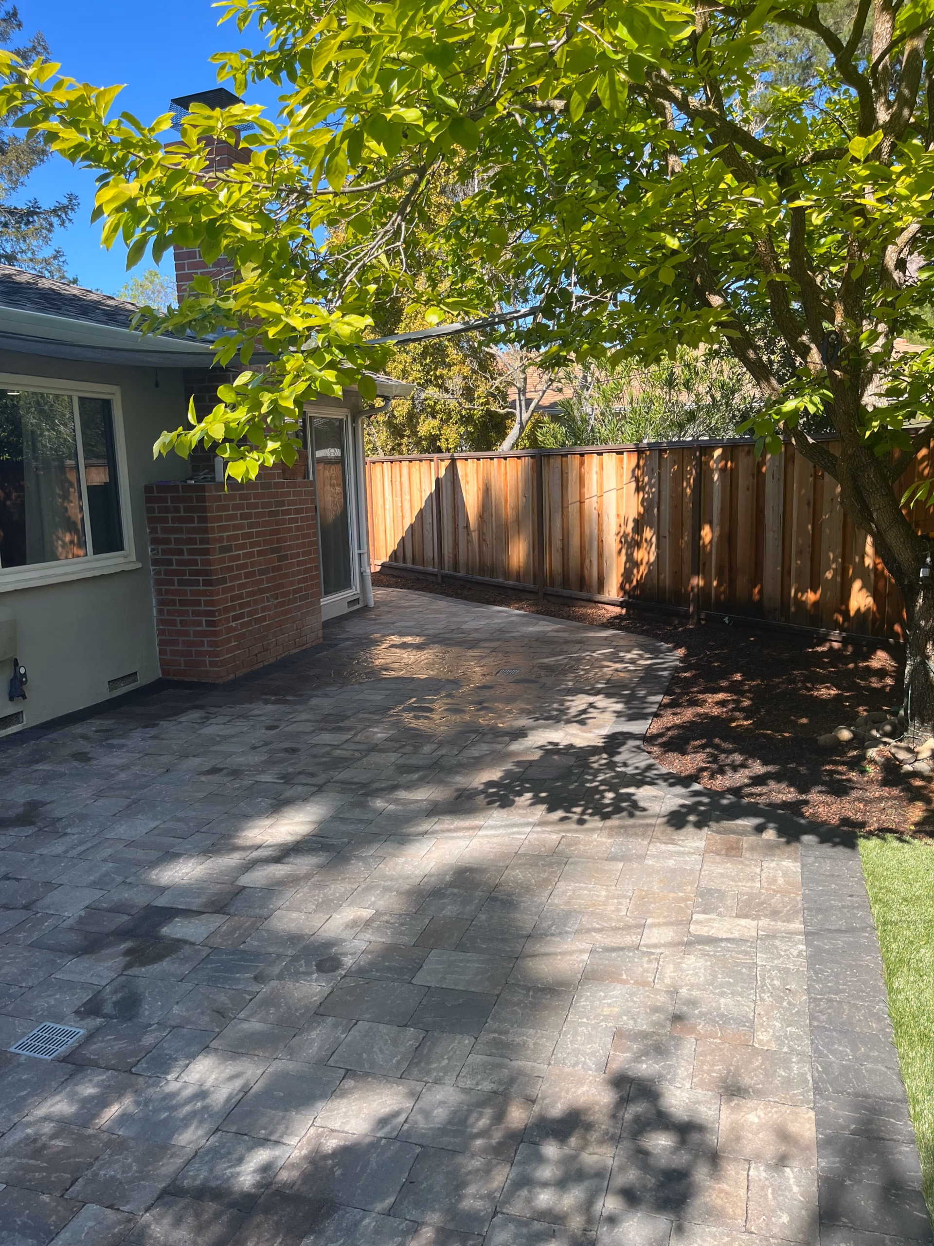 A paver terrace beneath a mature tree beside a brick-and-siding house, with a new wood fence, in Mountain View
