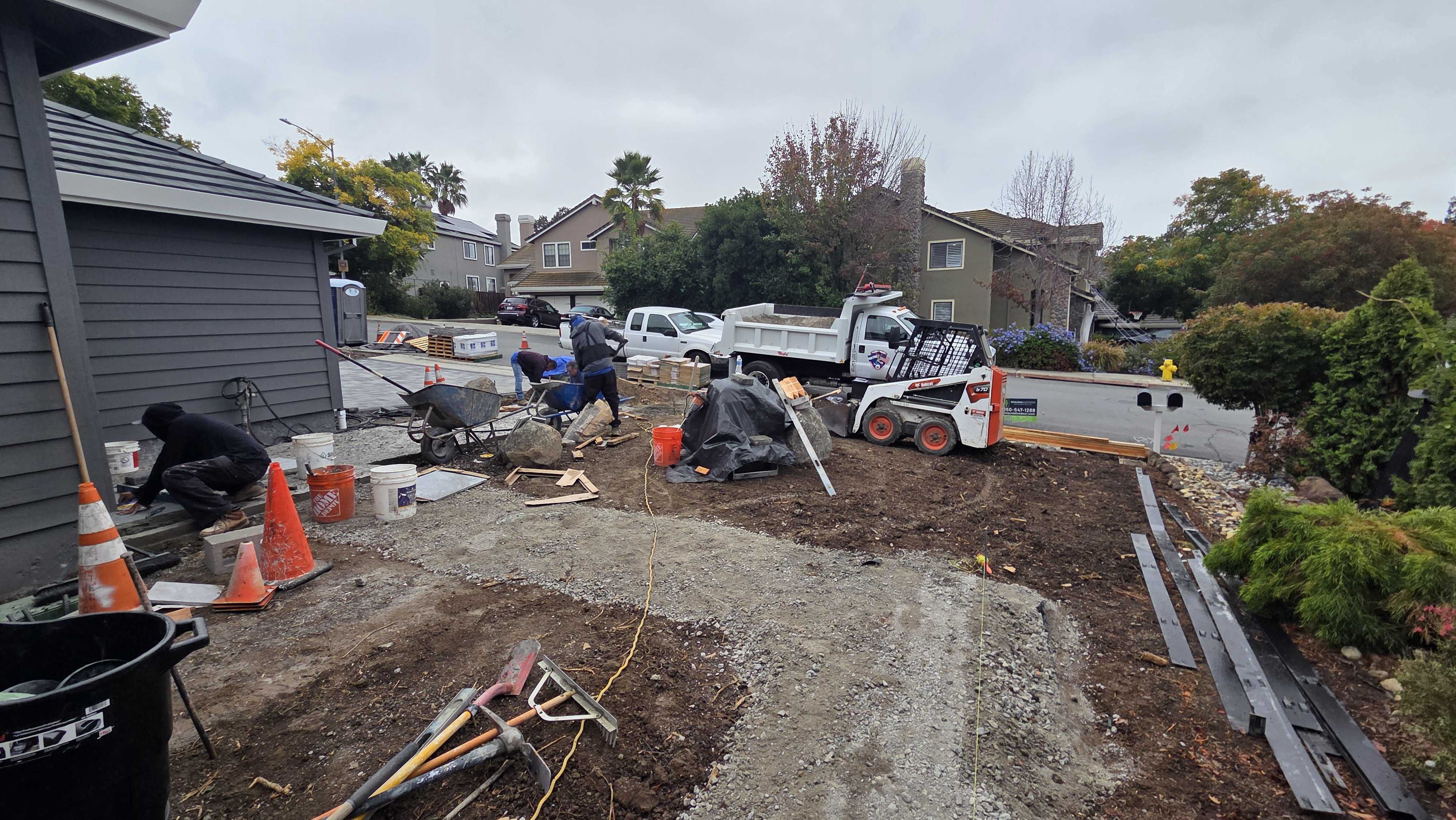Front-yard demolition and grading underway with a skid-steer, dump truck, and crew clearing the old yard.