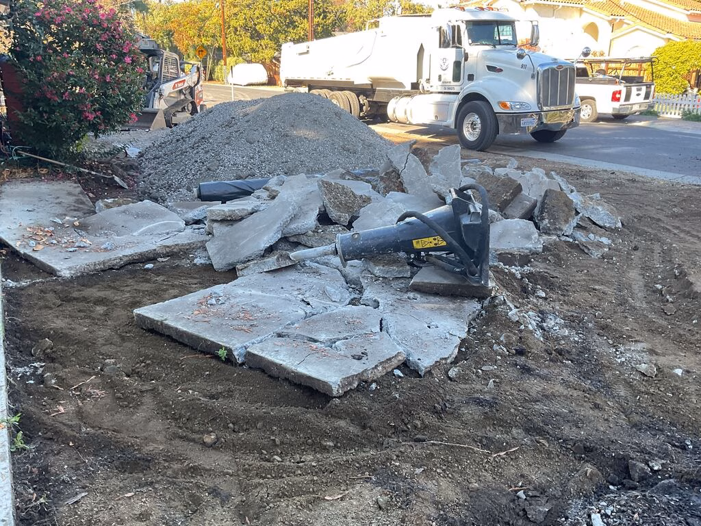 Broken concrete slab pieces from a driveway/sidewalk demolition with a handheld demolition hammer lying on top. An...