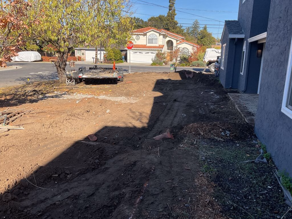 Residential front yard has been stripped of existing landscaping and shows exposed soil with equipment and a trailer...