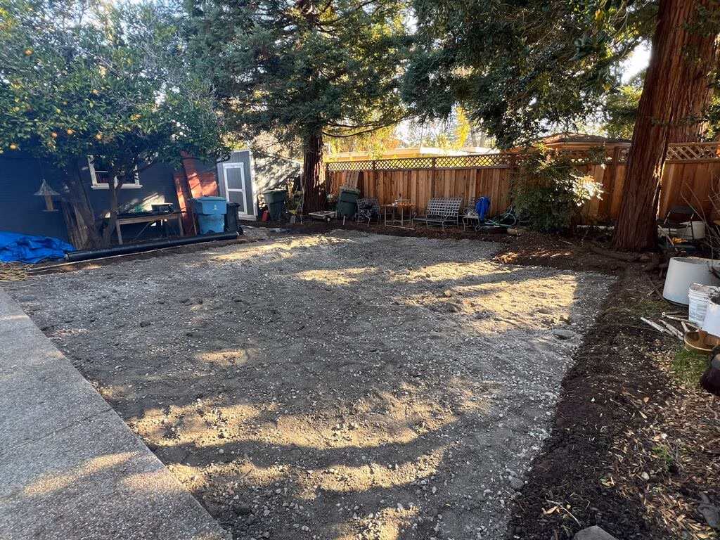 Cleared backyard with a compacted granular subbase and graded dirt surface, likely prepared for a future patio or turf...