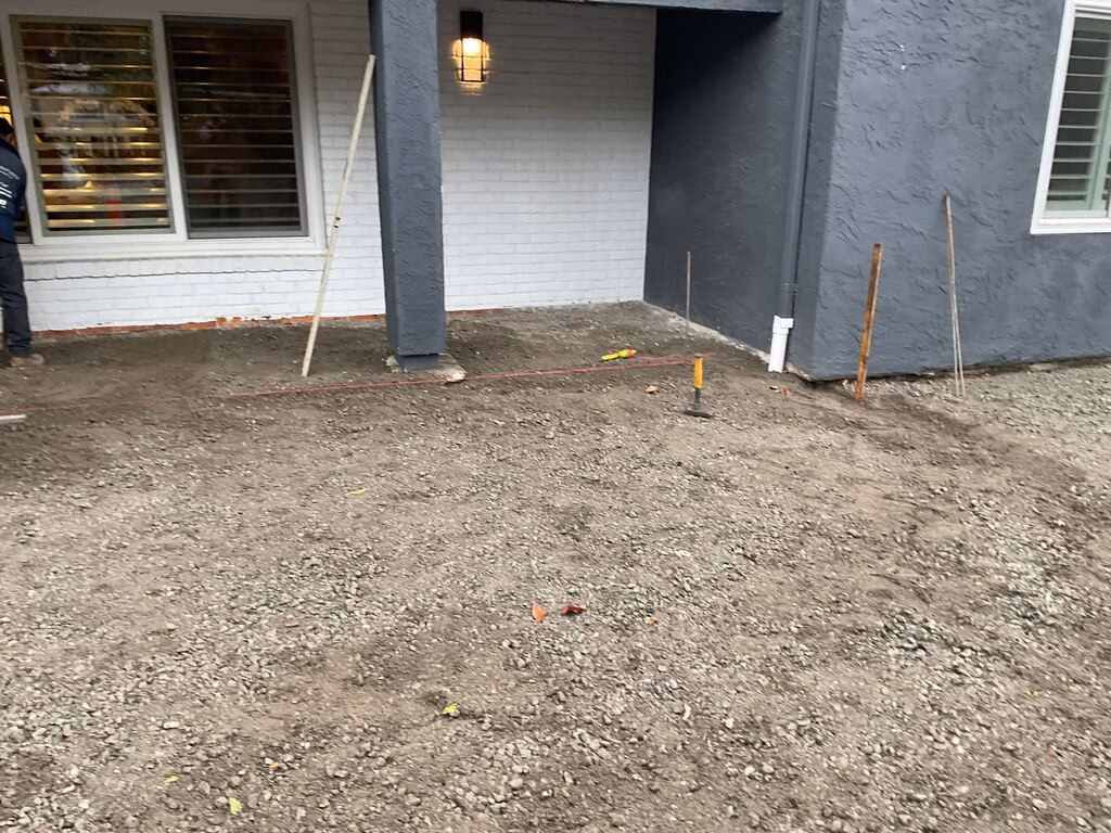 Prepped front porch and adjacent yard with a compacted crushed-rock base and grade stakes in place, ready for paver...