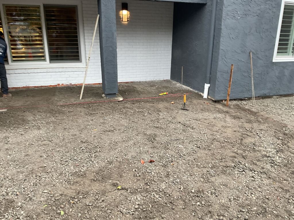 Prepped front porch and adjacent yard with a compacted crushed-rock base and grade stakes in place, ready for paver...