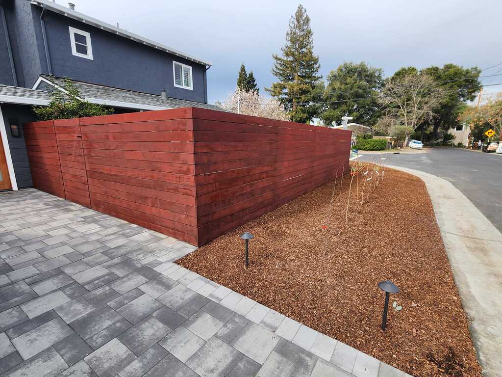Interlocking gray paver driveway beside a stained horizontal wood fence and a fresh bark planting bed.