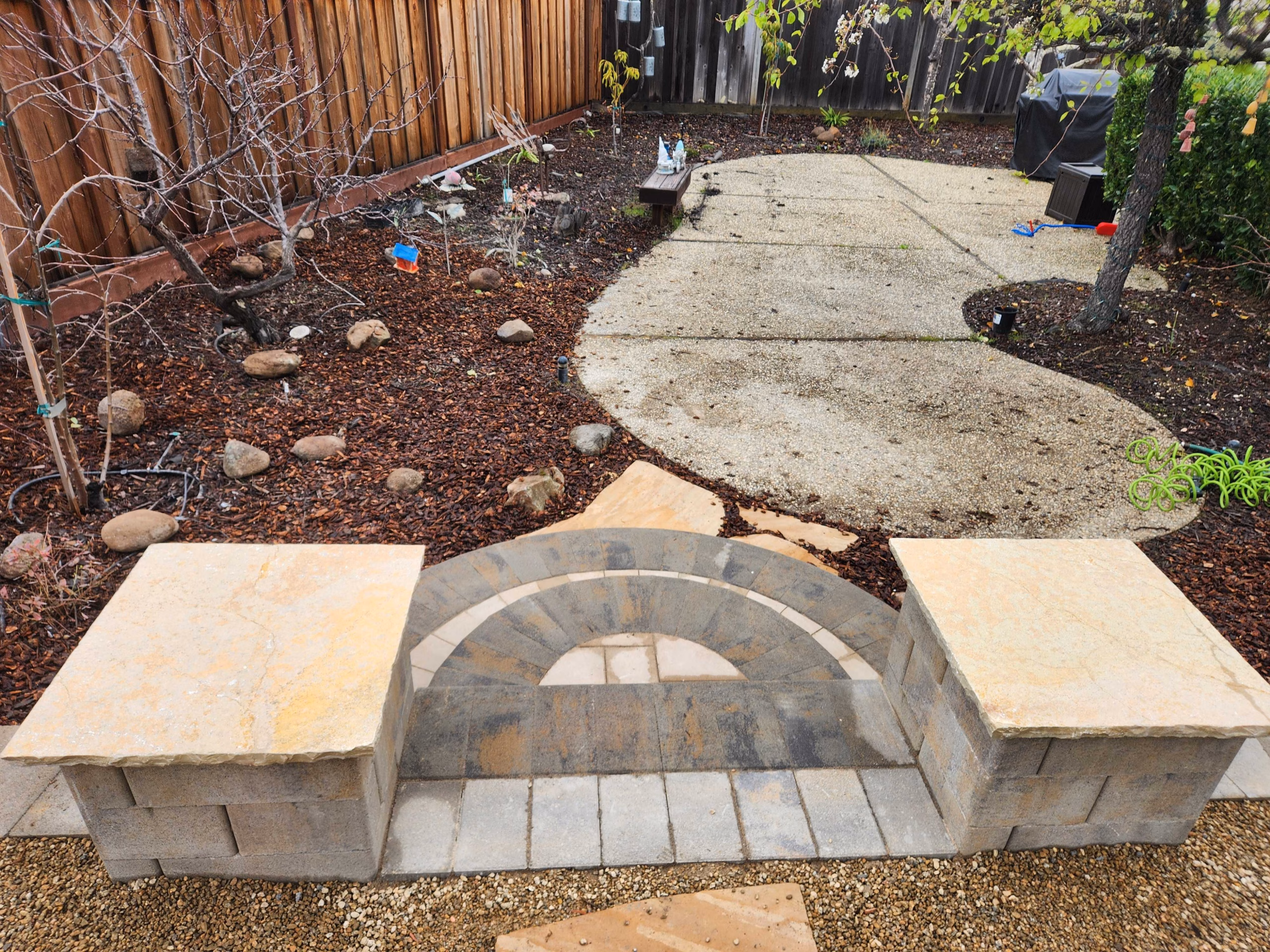 Block garden wall with an arched recess and flagstone-capped seat blocks, set on gravel beside an existing concrete path and a mulched planting bed, in Belmont, CA