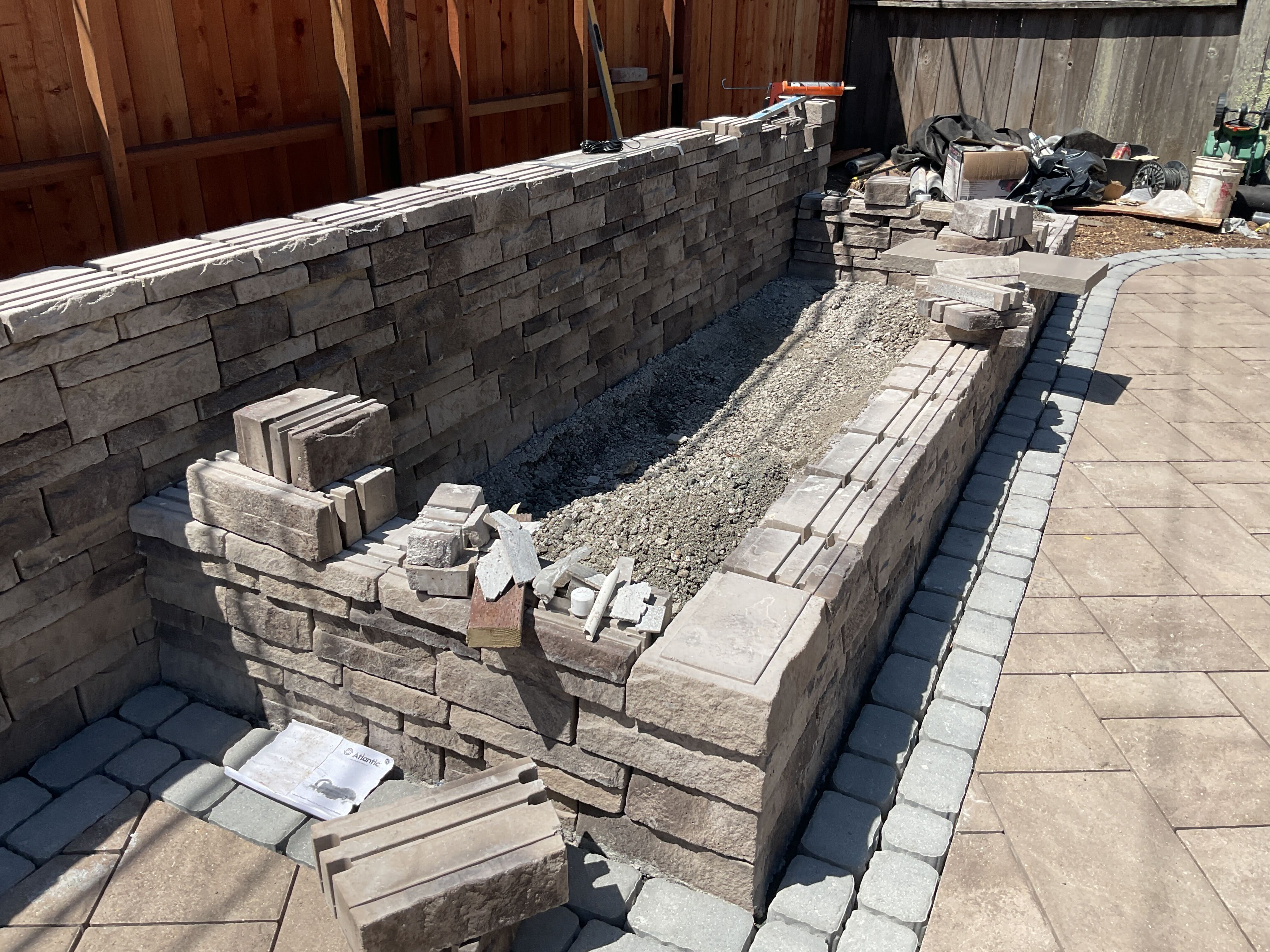 Stacked-stone seat wall mid-build: open gravel core with loose veneer blocks staged on the cap and finished pavers in front.