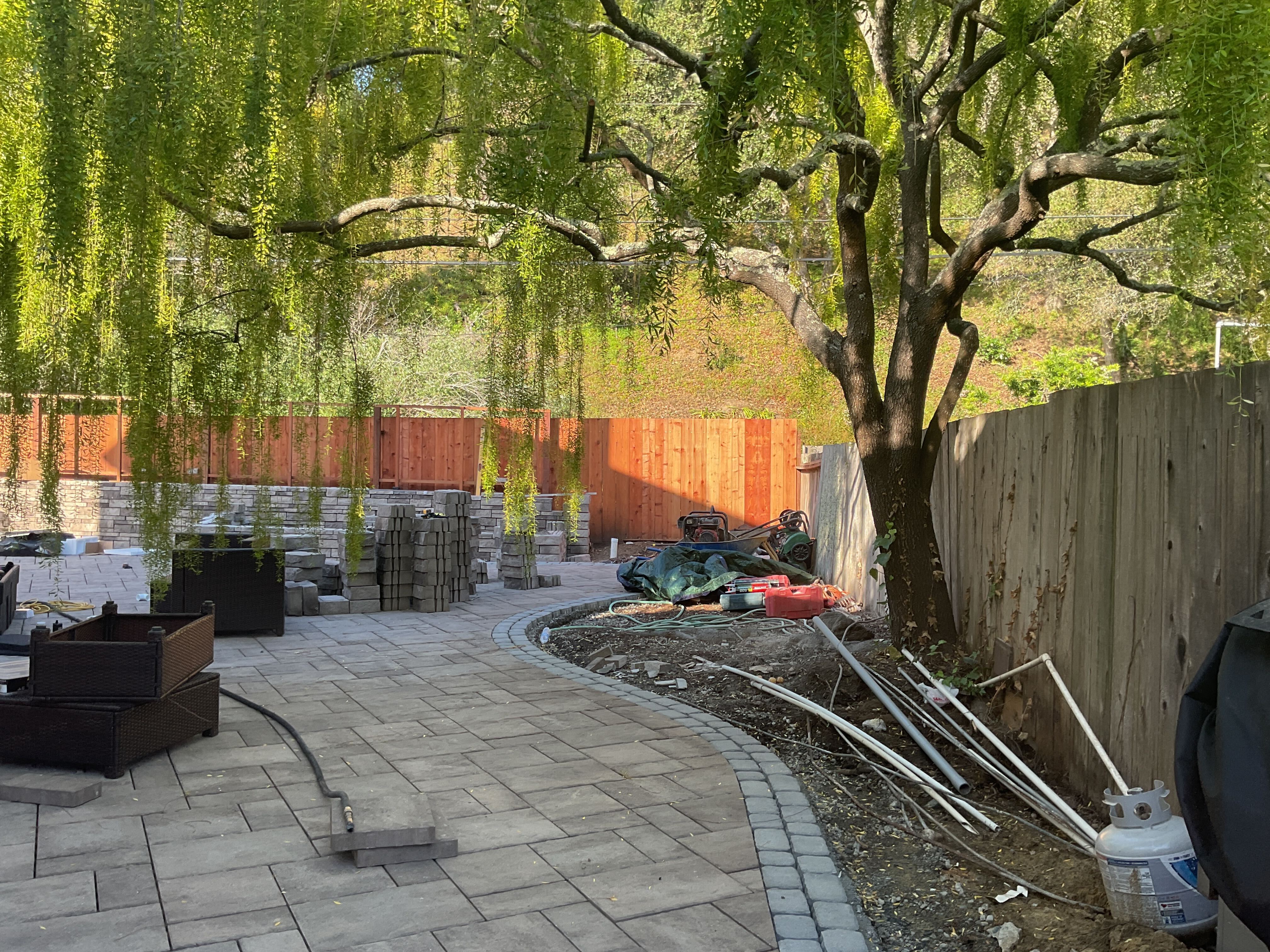 Paver field mid-lay with the curved cobble border being set, willow and cedar fence behind, raw-dirt planting strip not...
