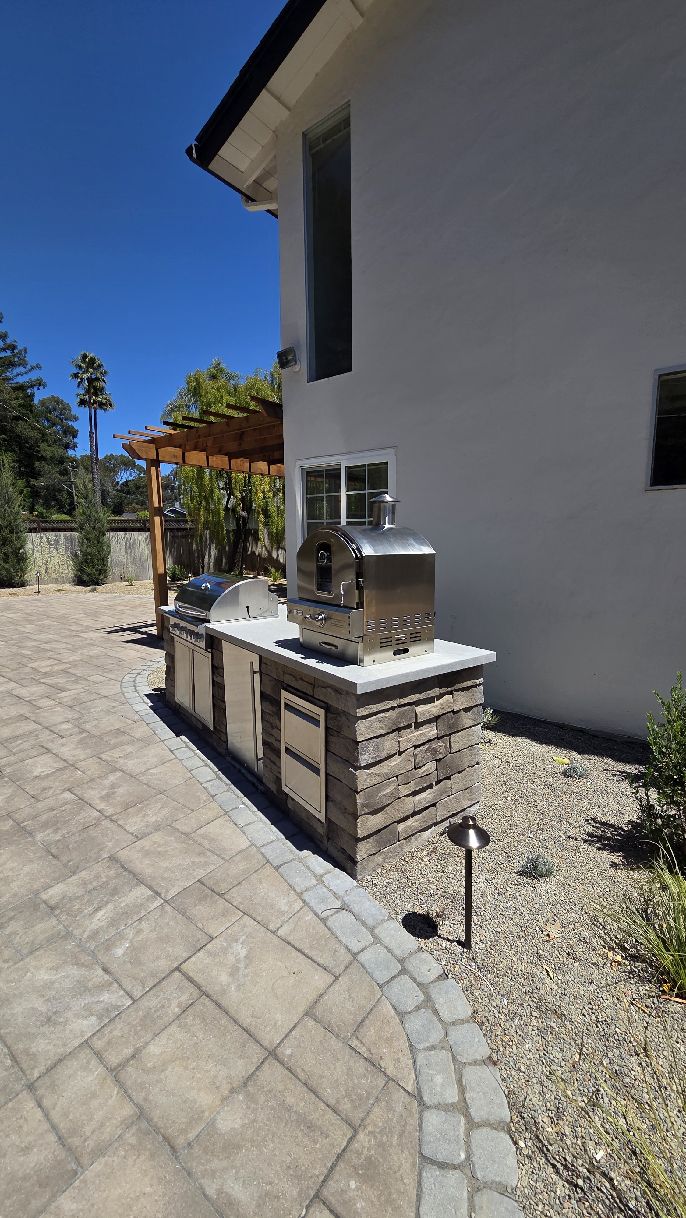 Stone-clad outdoor kitchen island with a stainless grill and countertop pizza oven and a white concrete cap, on a...