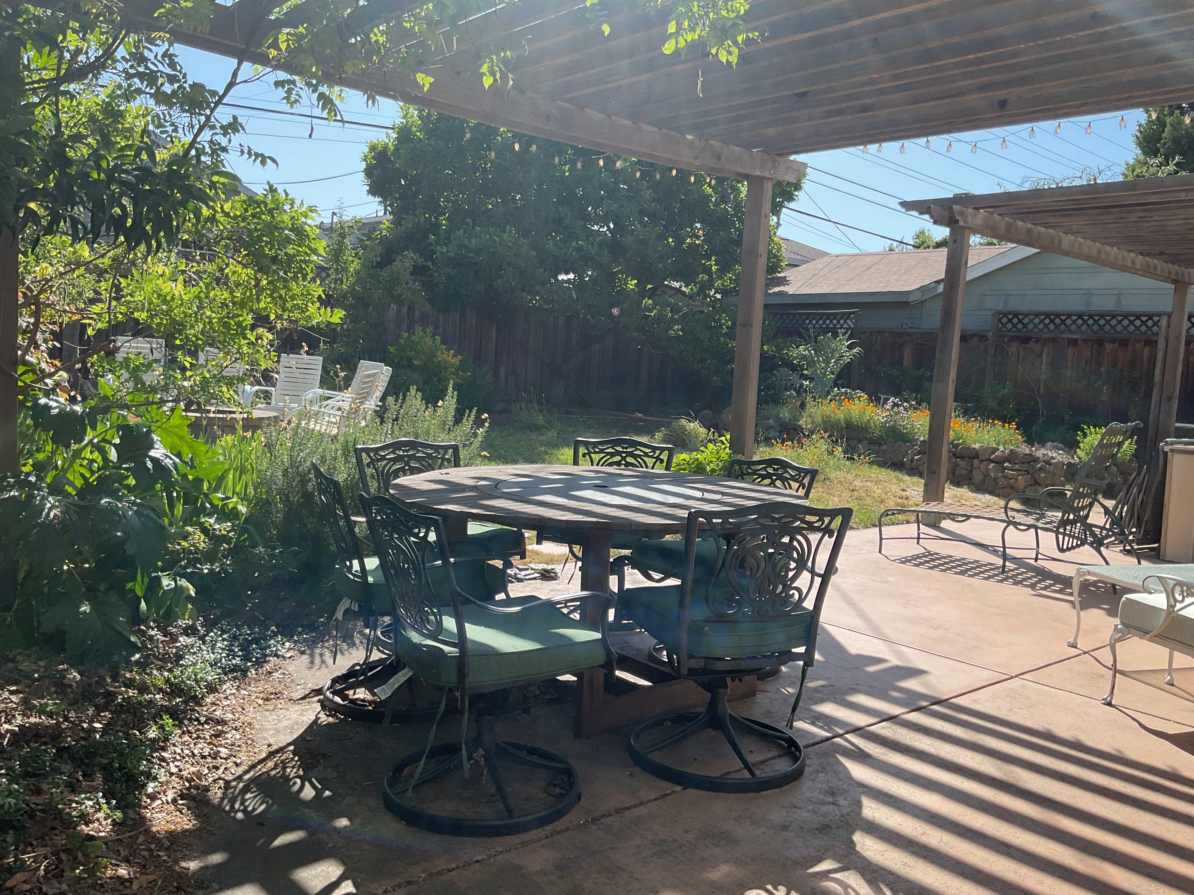 Shaded concrete patio under the wood pergola with string lights, a dining set, and planting beds along the stone wall.