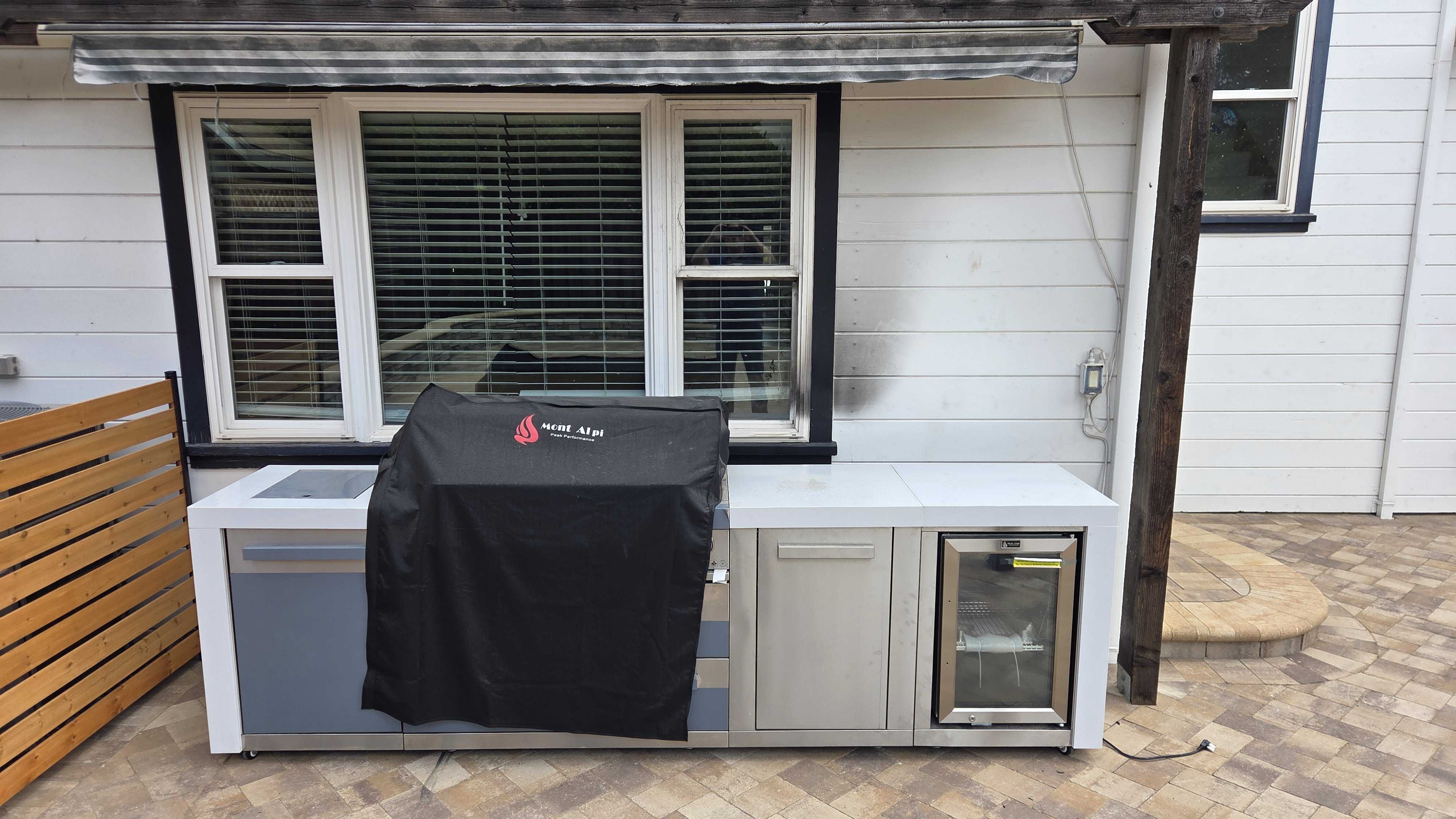 Built-in outdoor kitchen installed against the house exterior, featuring a covered grill, stainless-steel cabinetry, a...