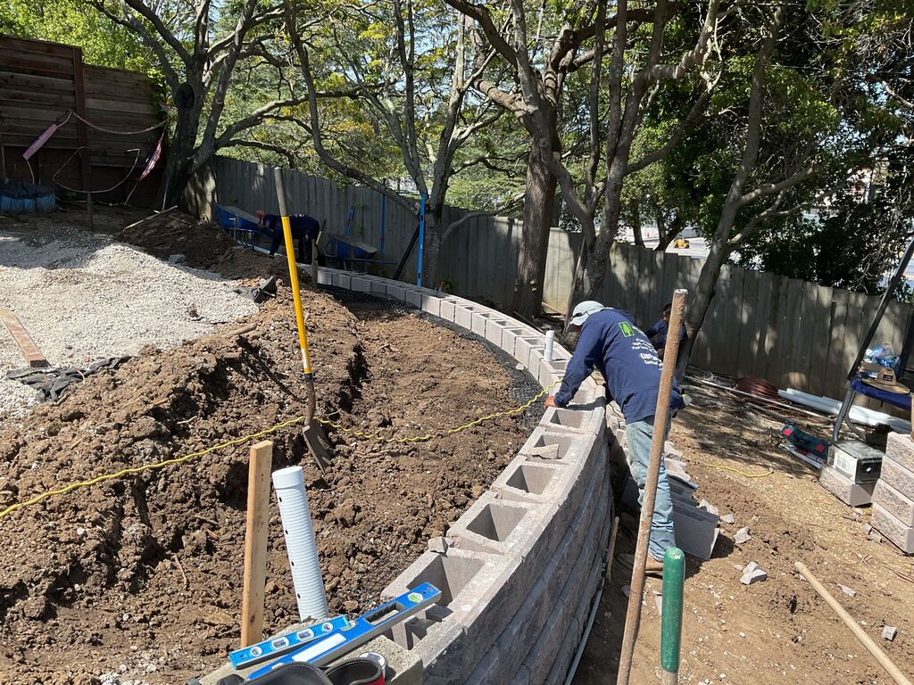Crew laying the curved segmental retaining wall course by course, with drainage pipe and gravel backfill behind the block.