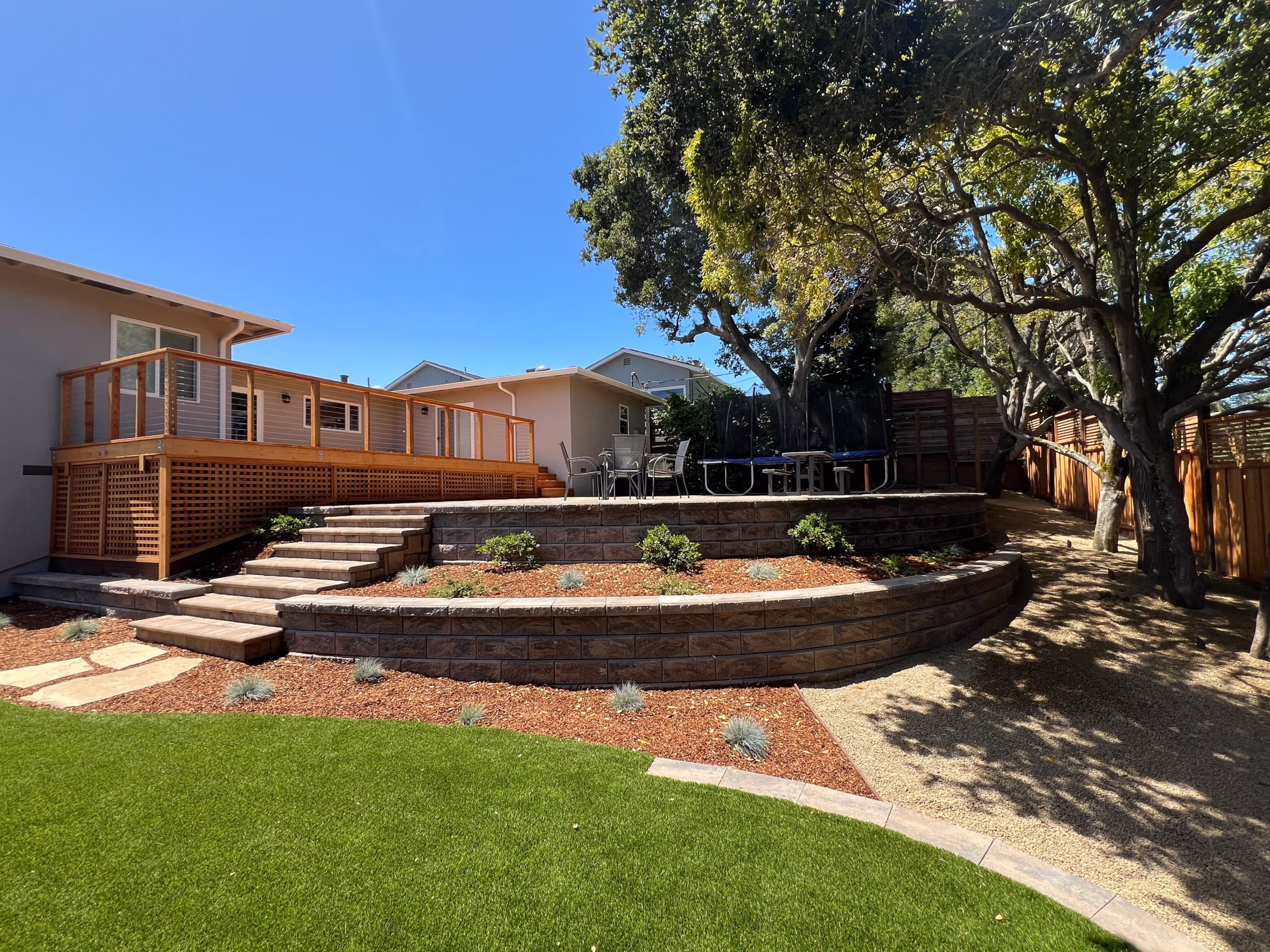 Terraced backyard with curved segmental retaining walls, paver steps to a redwood deck, artificial turf, and planting beds.