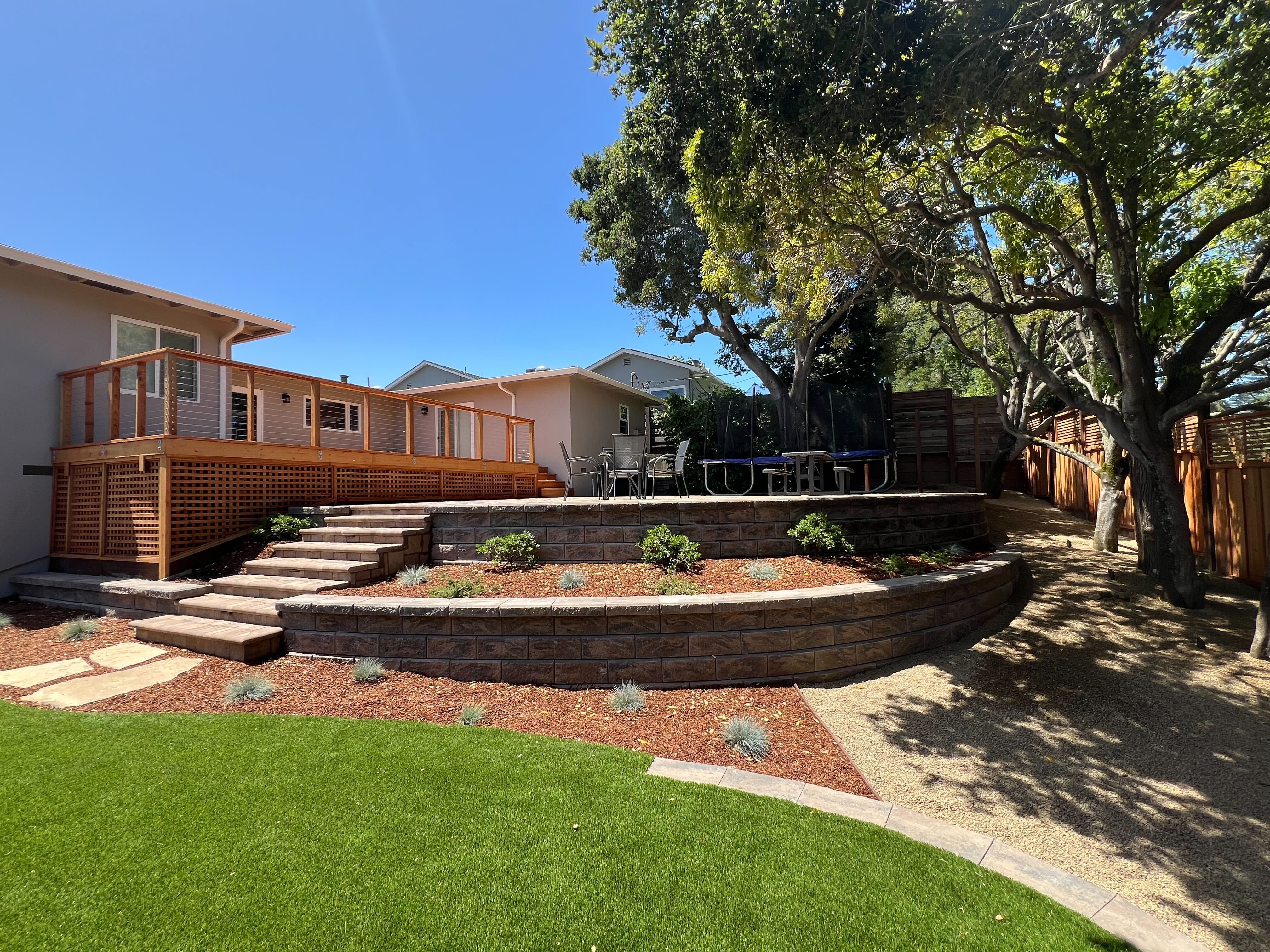 Terraced backyard with curved segmental retaining walls, paver steps to a redwood deck, artificial turf, and planting beds.