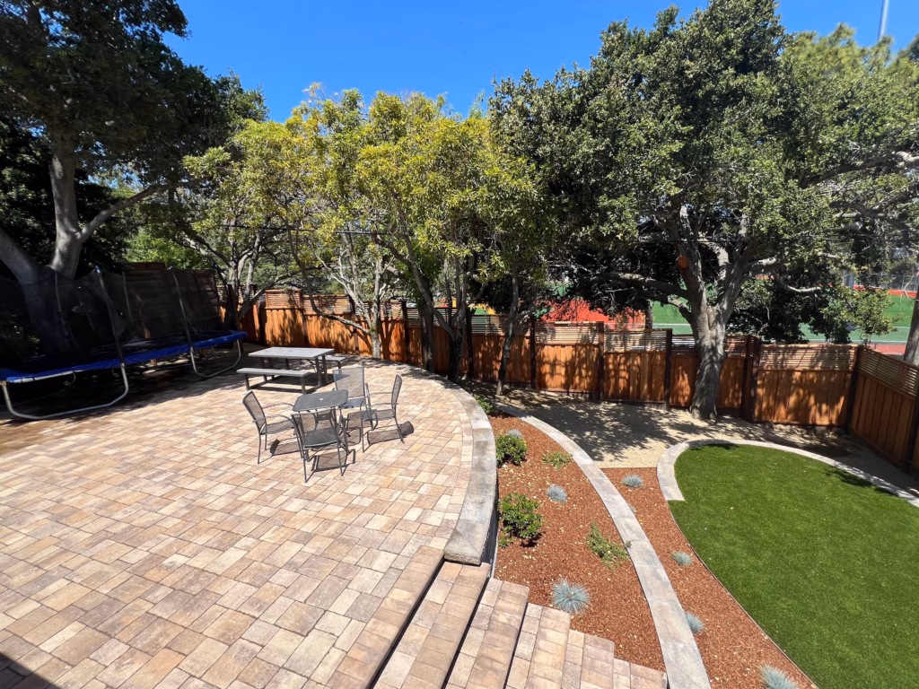 After — Curved Calstone paver patio with seating, a low capped border wall, planting beds, and an artificial-turf lawn along the fence.