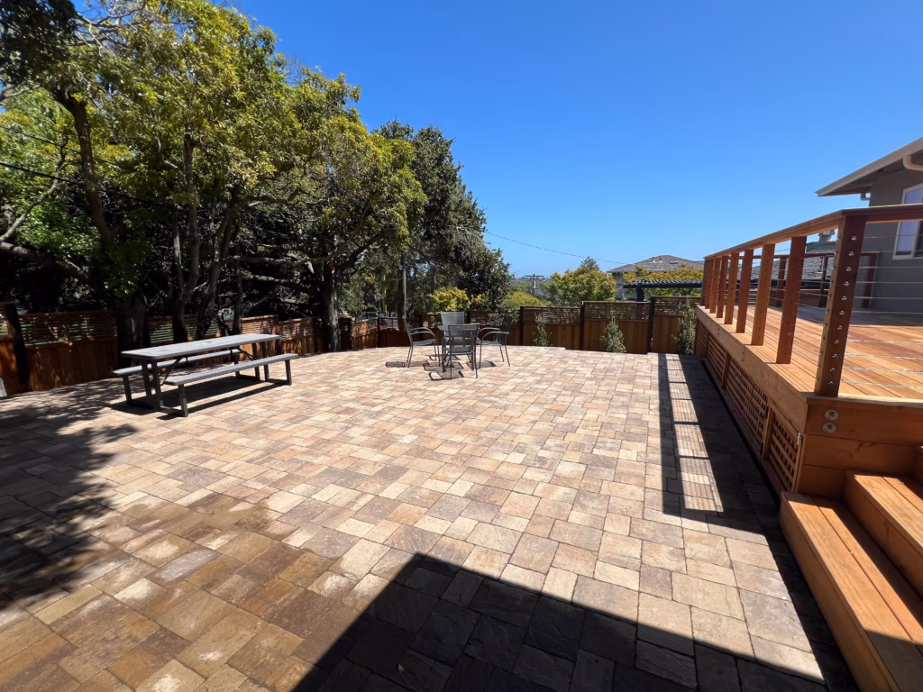 Wide paver patio bordered by a dark contrasting band, beside the raised deck with cable railing and hill views.