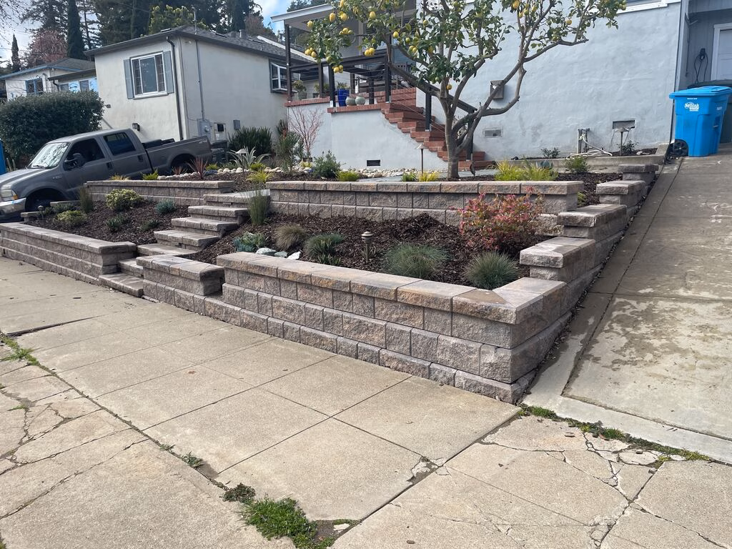 After — Finished front yard with a tiered, capped segmental retaining wall, steps up from the sidewalk, and new planting beds.