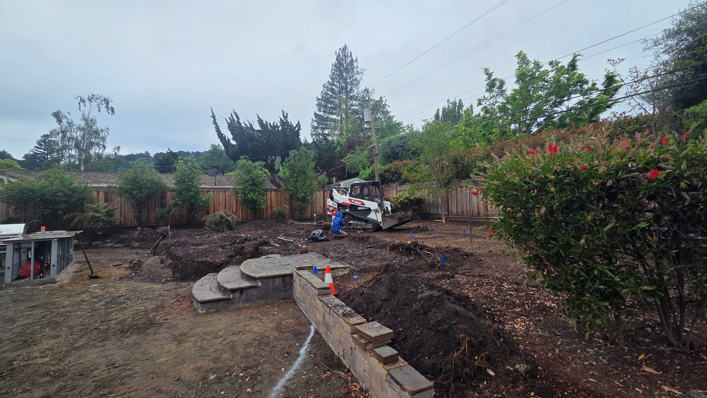 Skid-steer grading around the partially built retaining wall and circular paver landing.