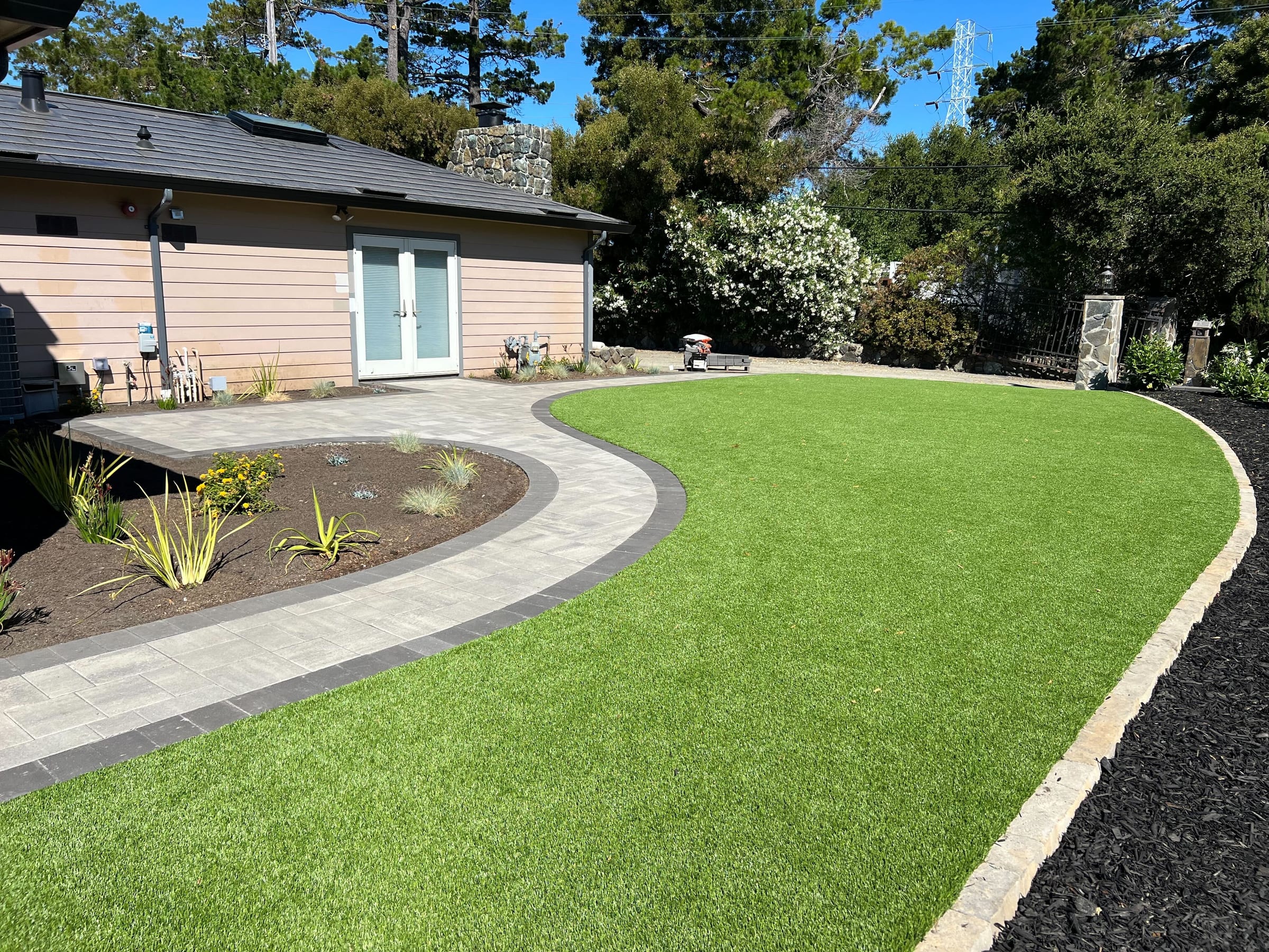 Curved paver walkway with a dark border band separating the turf lawn from a new planting bed near the stone pillar.
