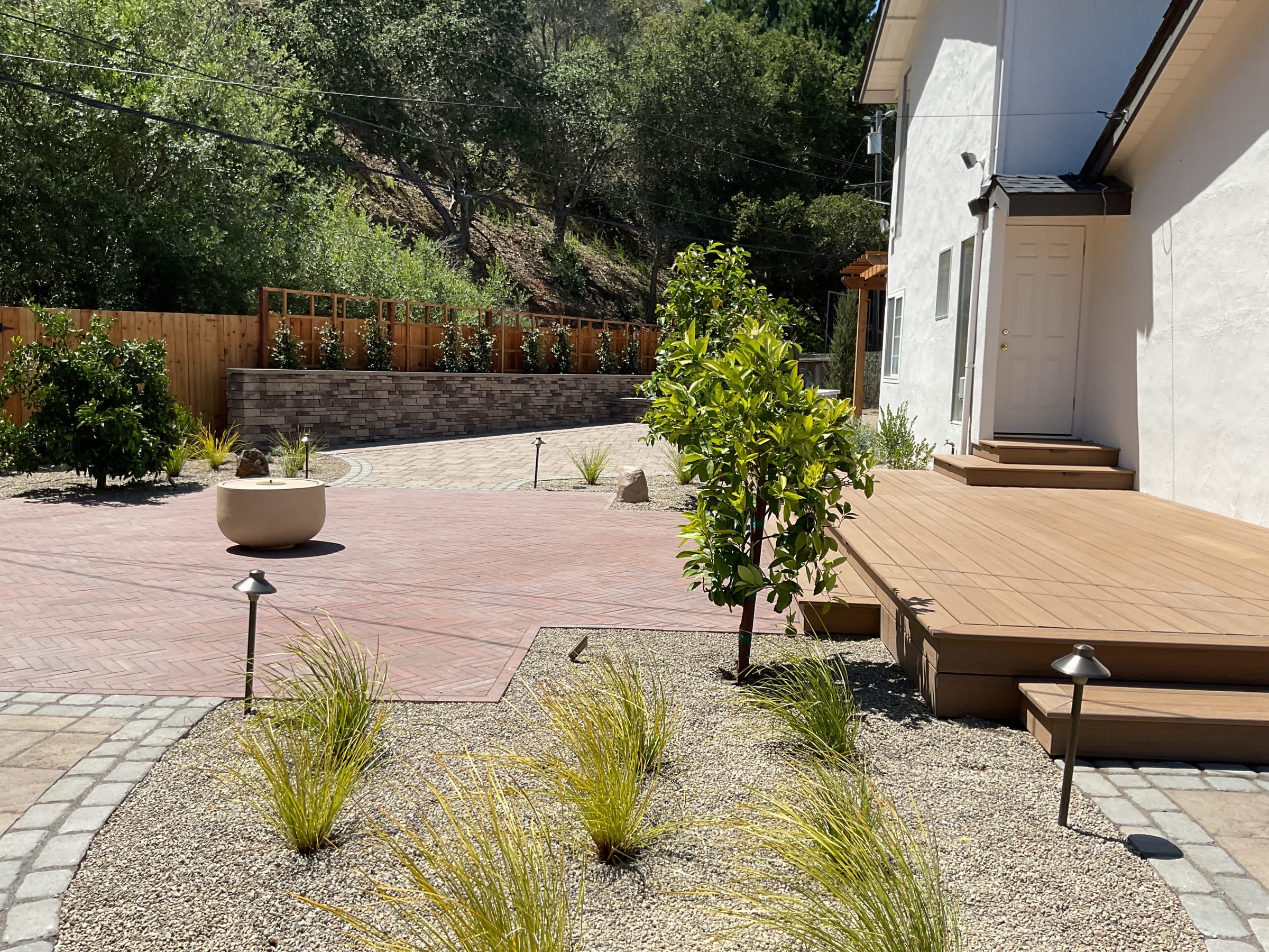 Finished paver patio beside the composite deck with gravel planting beds, path lights, and a stone seat wall beyond.
