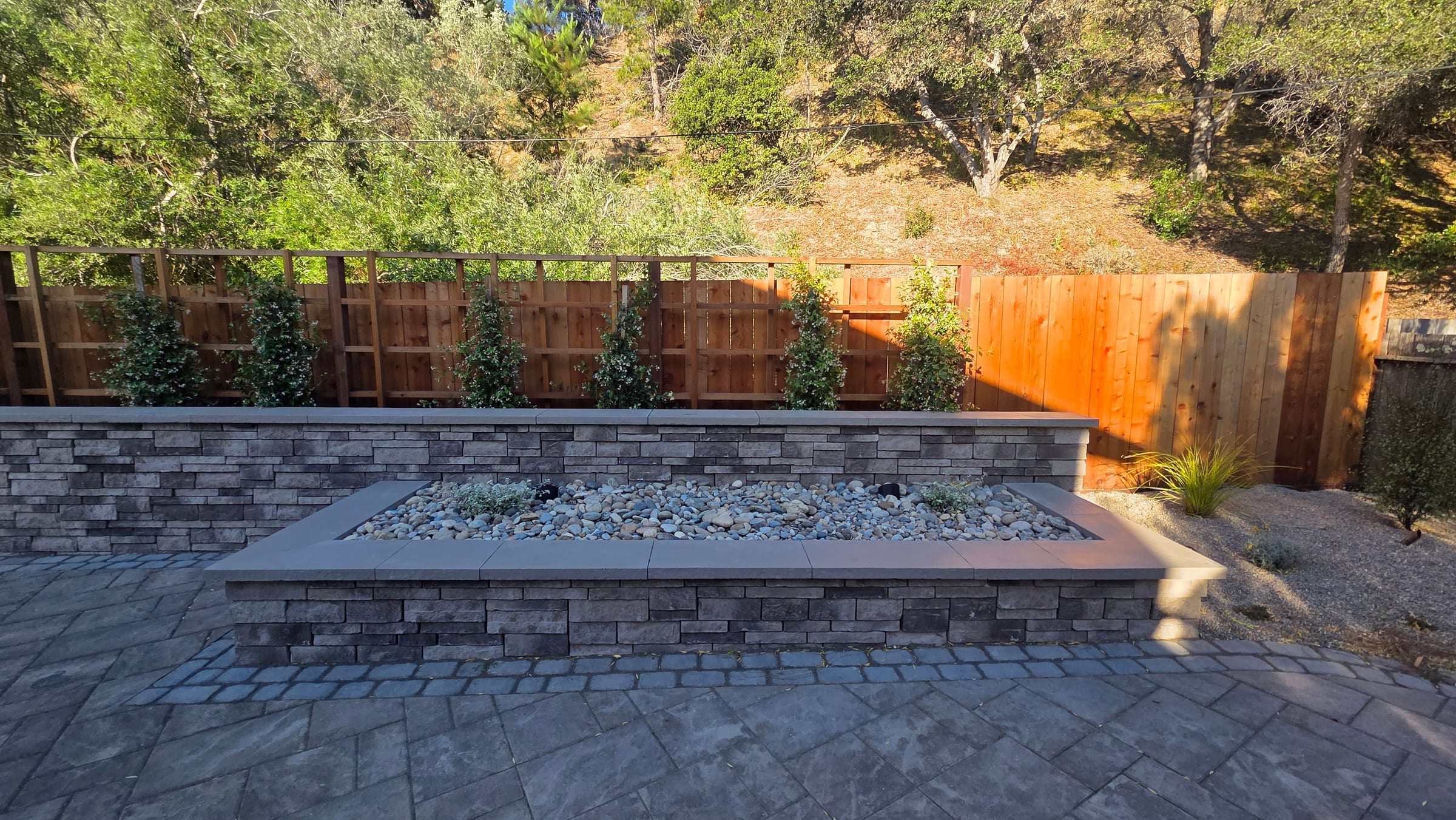 A capped stacked-stone planter filled with river rock sits on the paver patio in front of a cedar privacy fence.