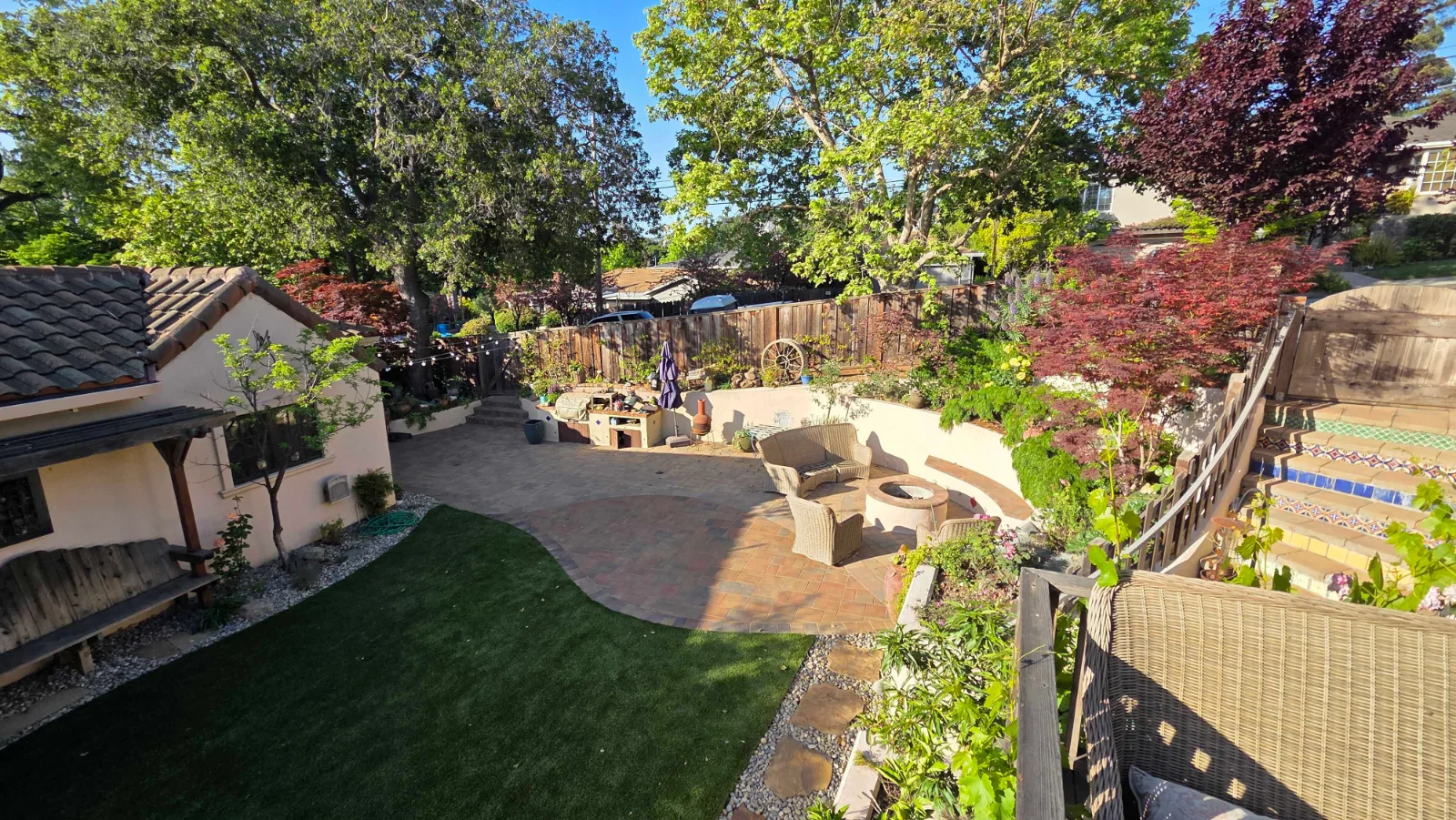 An elevated view of a finished backyard: a curved paver patio with a round fire pit and wicker armchairs, a turf lawn, and mature Japanese maples along a stained wood fence