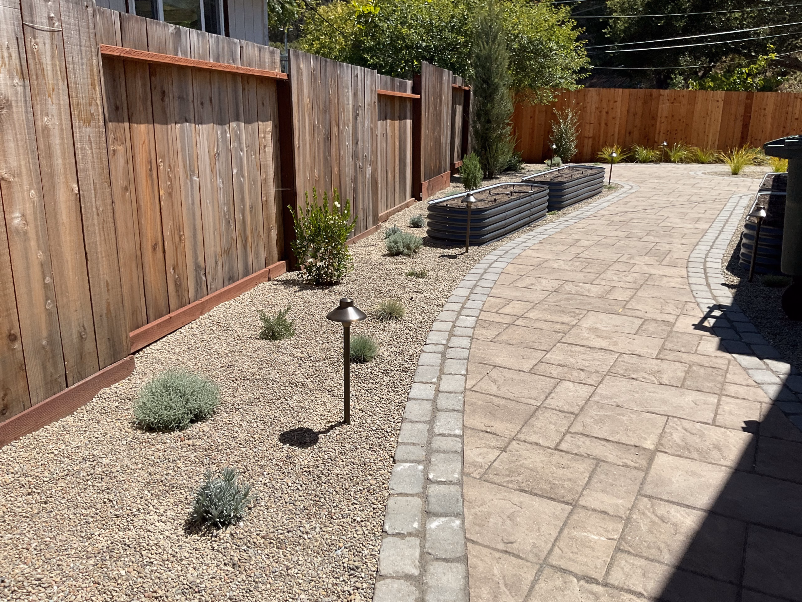 Paver walkway with a brass path light, cobble border, gravel planting beds and raised garden beds along the fence.