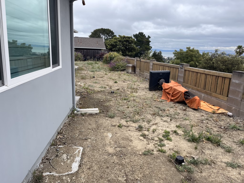 Before — bare dirt and weeds along the back of a Burlingame house, fence and trees beyond