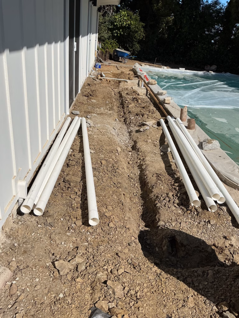 Lengths of white drain pipe laid beside a trench along a board-and-batten building, pool under a protective cover