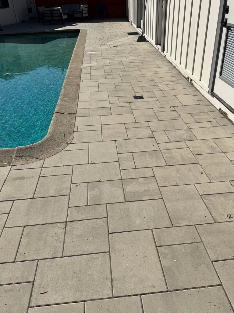 Paver walk between a pool and a white board-and-batten building, with a seating area at the far end